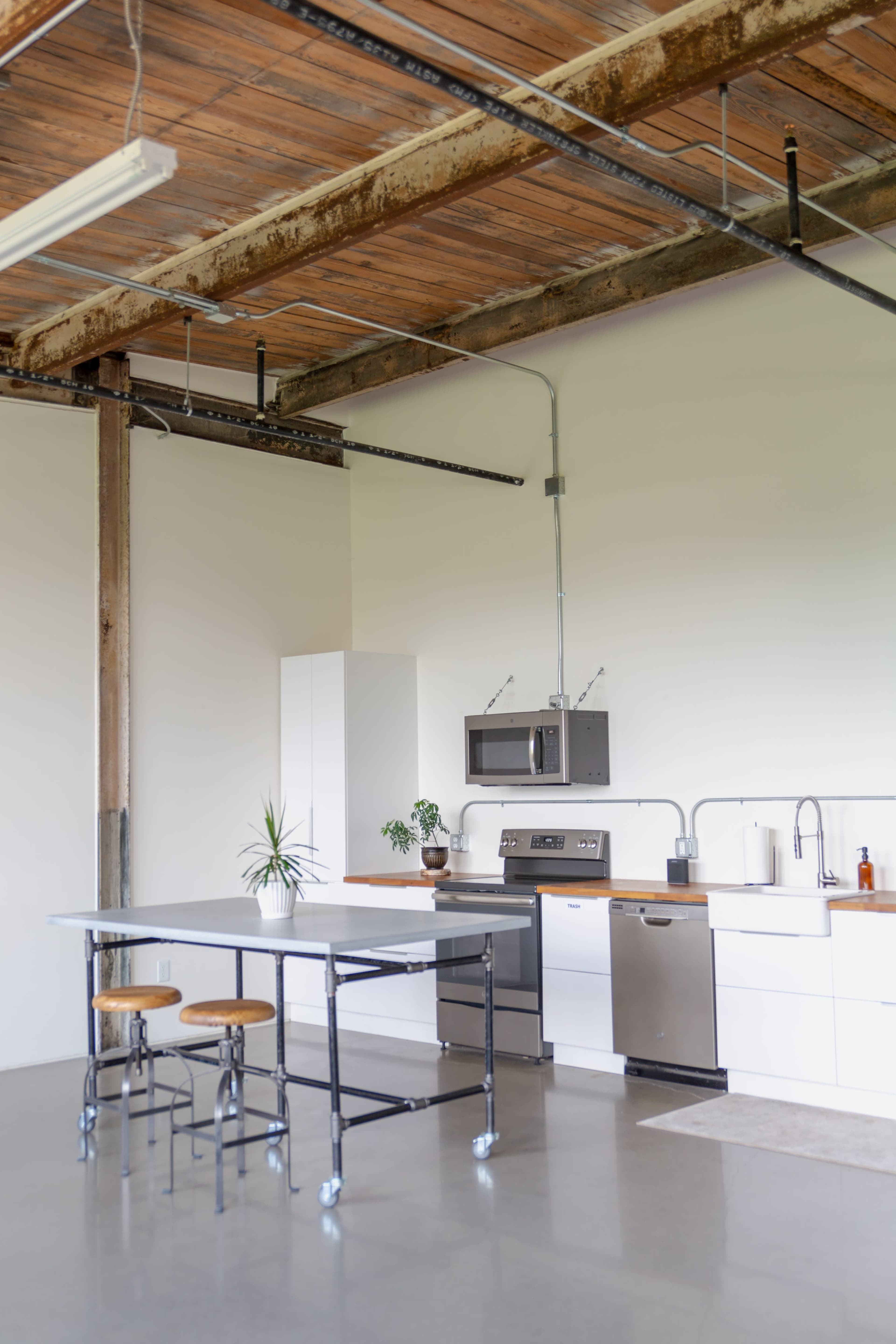 A modern kitchen with a metal table and stools, stainless steel appliances, and a minimalist design featuring white cabinets and a wooden countertop.