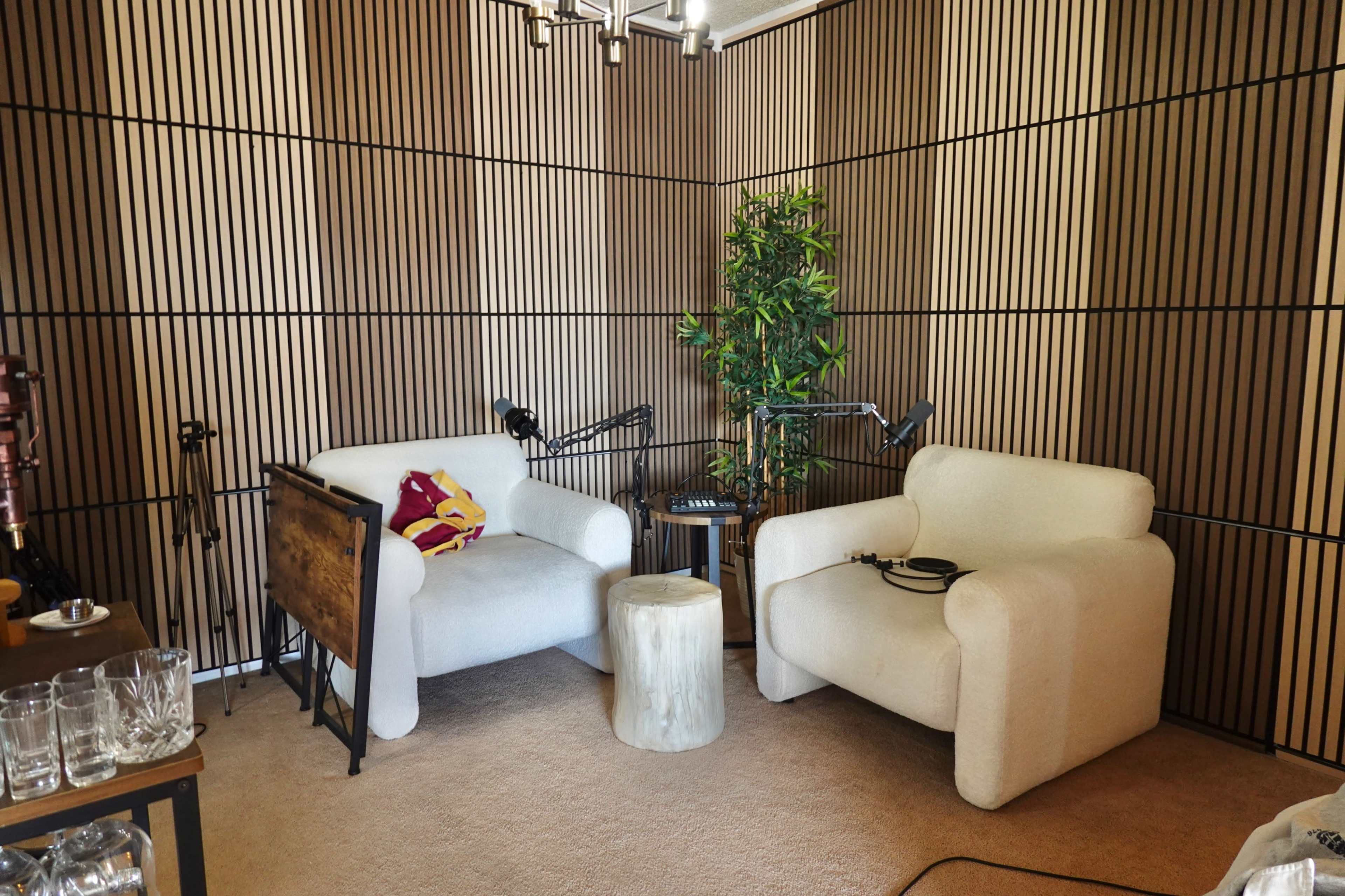 The image shows a cozy recording studio with two white chairs, a small round table, and audio equipment, all set against a wood-paneled backdrop.