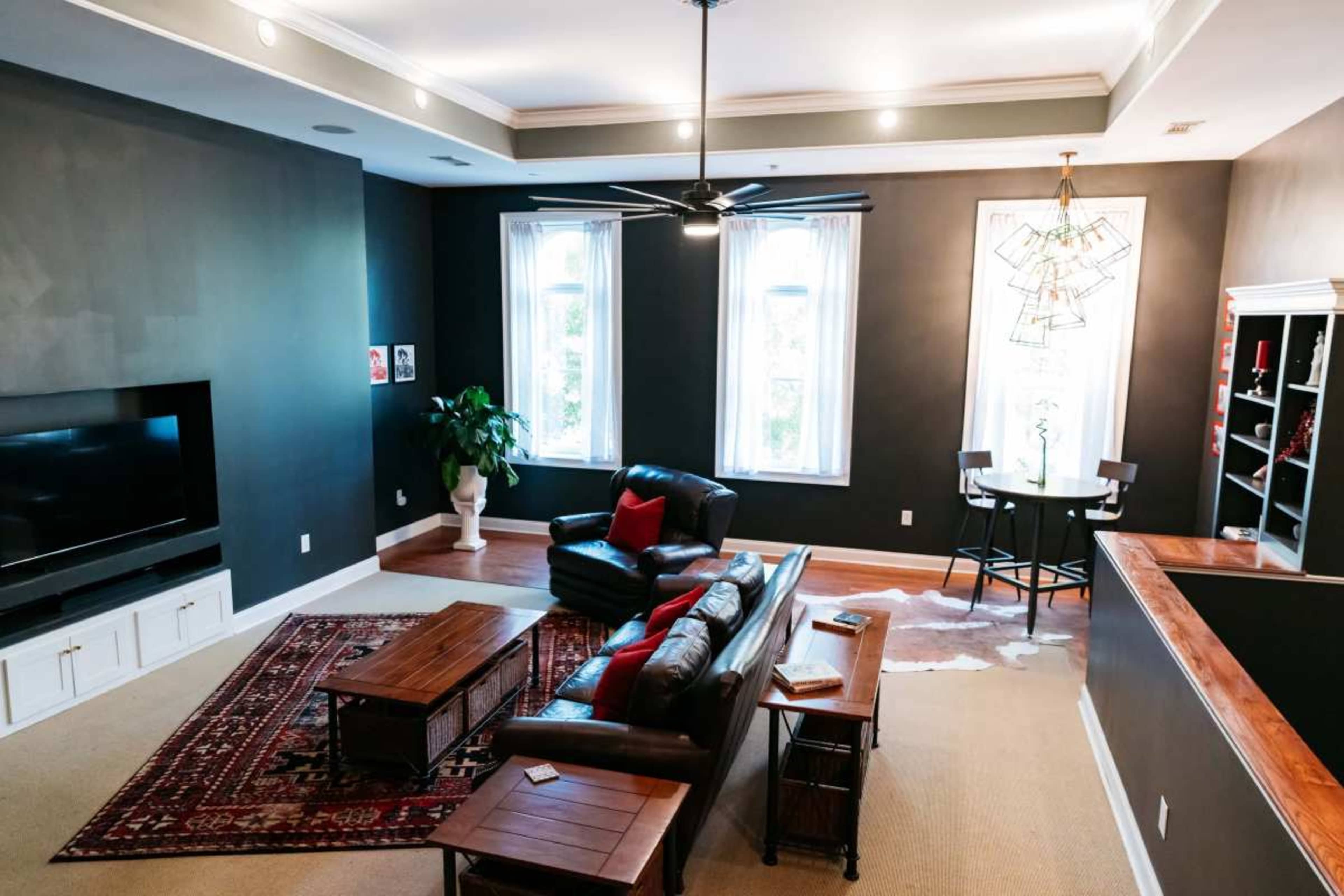 The spacious living room features a dark wall color, a leather sofa, a coffee table, and two windows with sheer curtains.