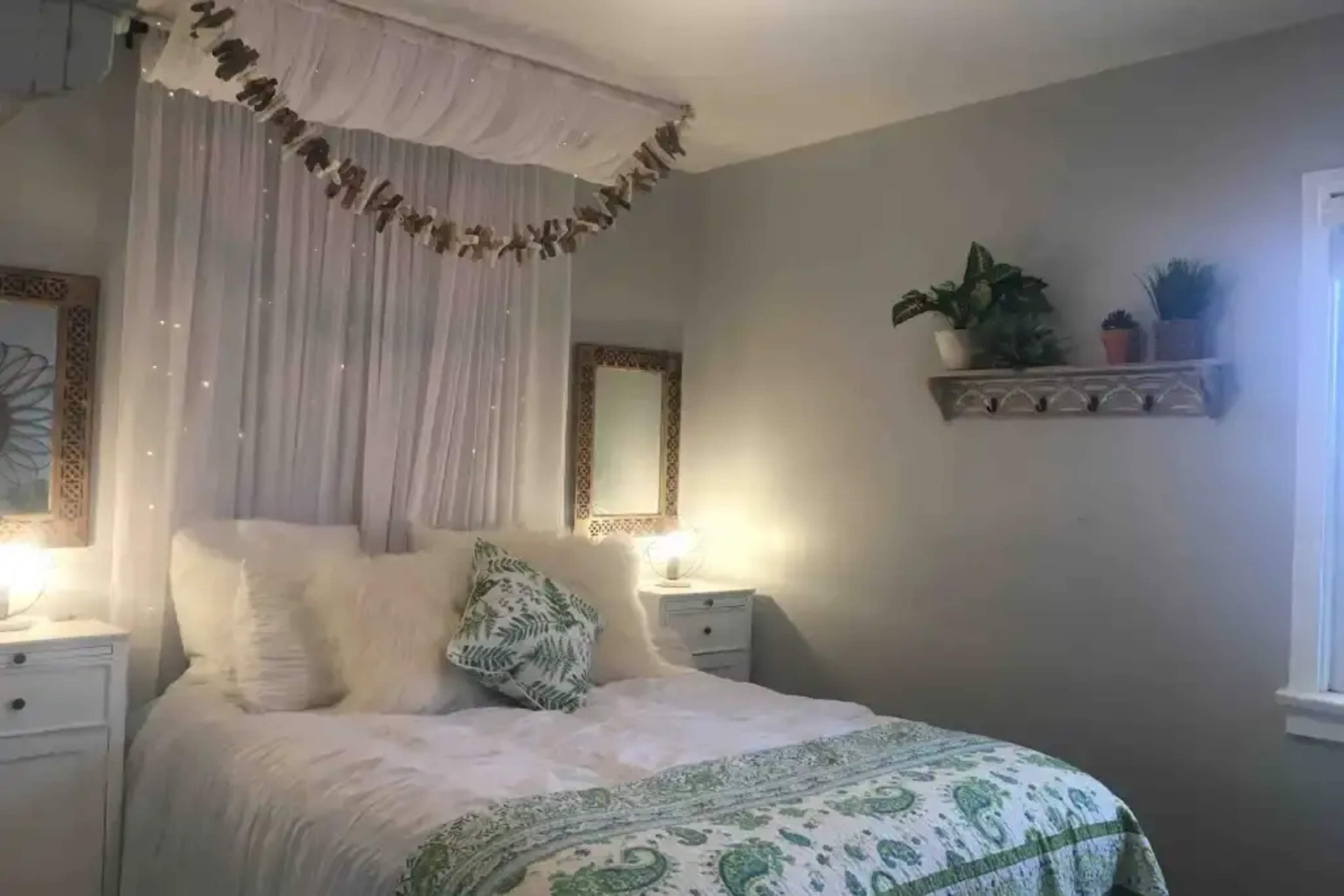 A well-decorated bedroom features a bed with white linens, a patterned throw, and plants on a shelf, complemented by string lights and mirrors on the walls.