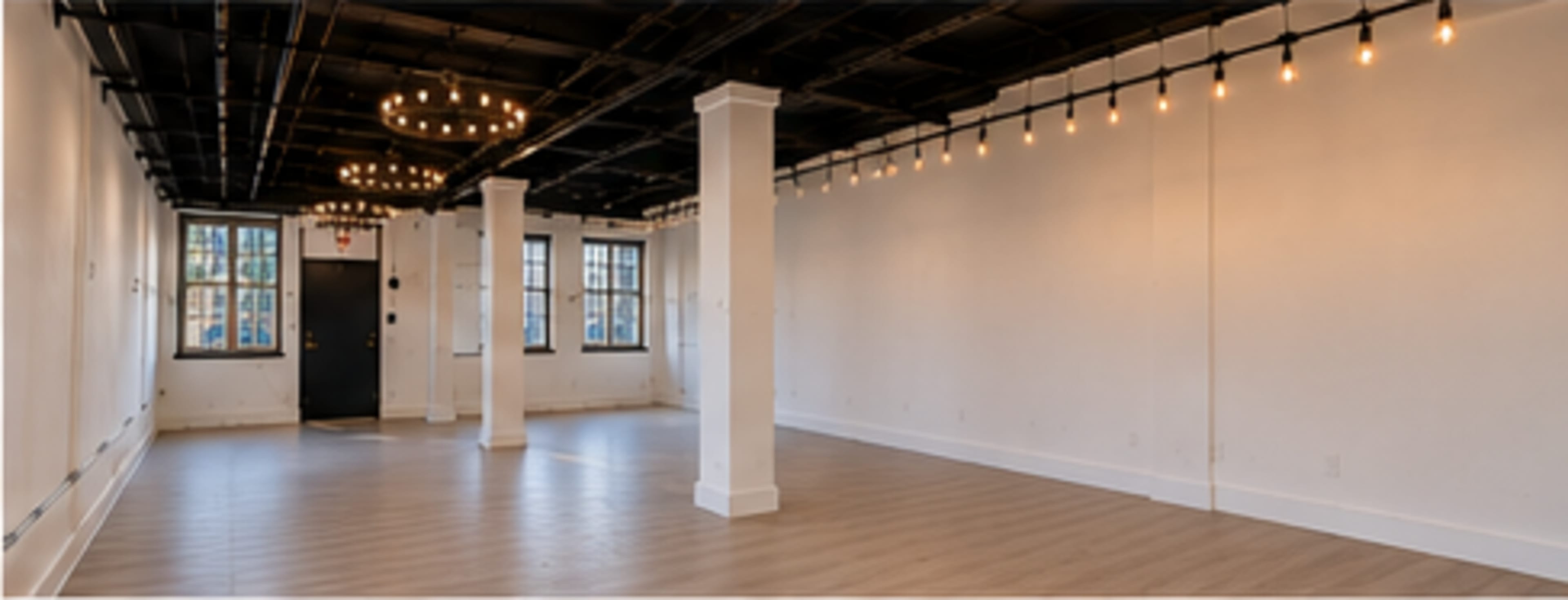 The image depicts an empty indoor space with wooden flooring, white walls, and exposed black ceiling beams featuring hanging light fixtures.
