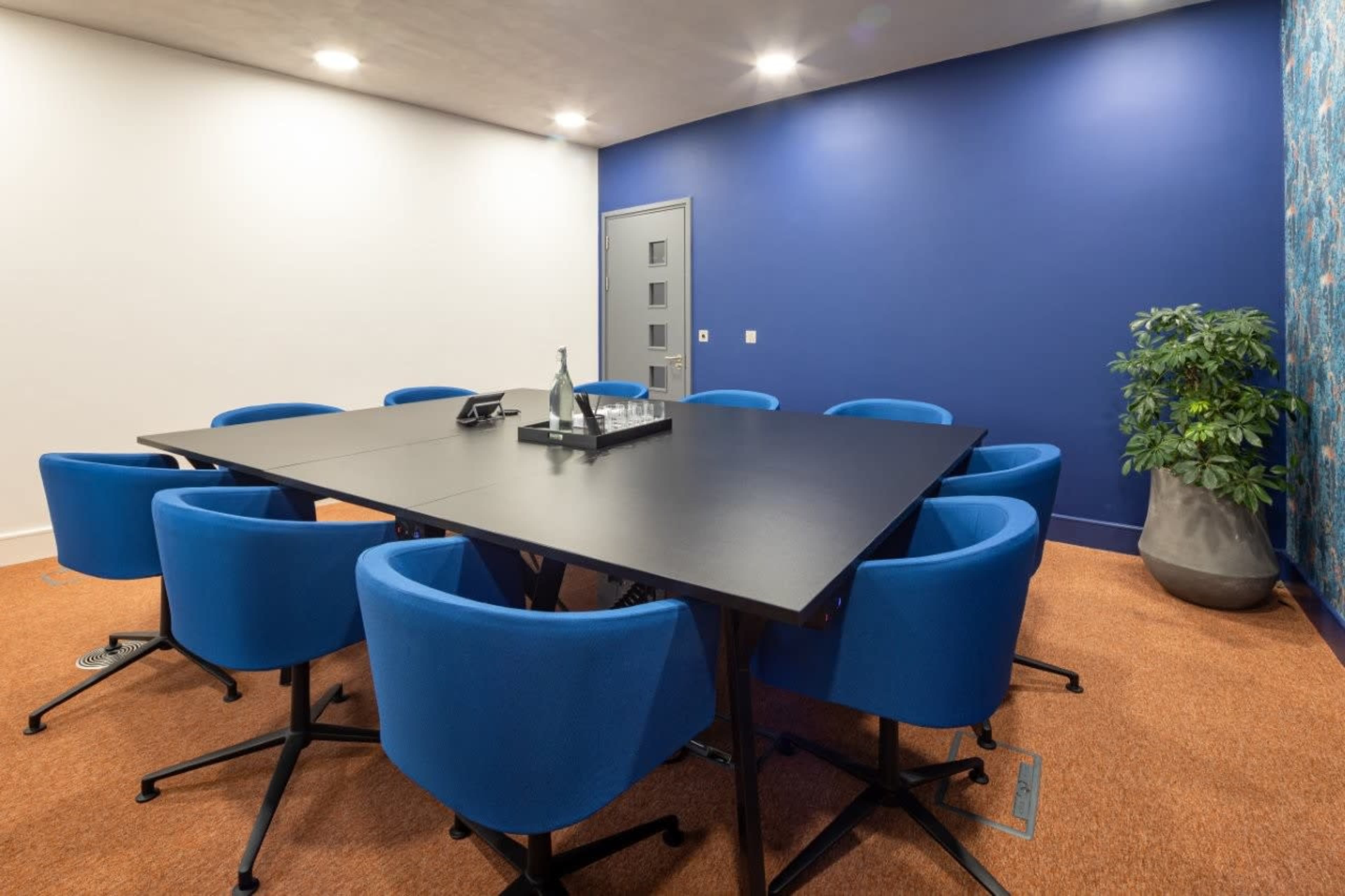 A modern conference room features a large black table surrounded by blue chairs, with a wall adorned in blue and floral design.