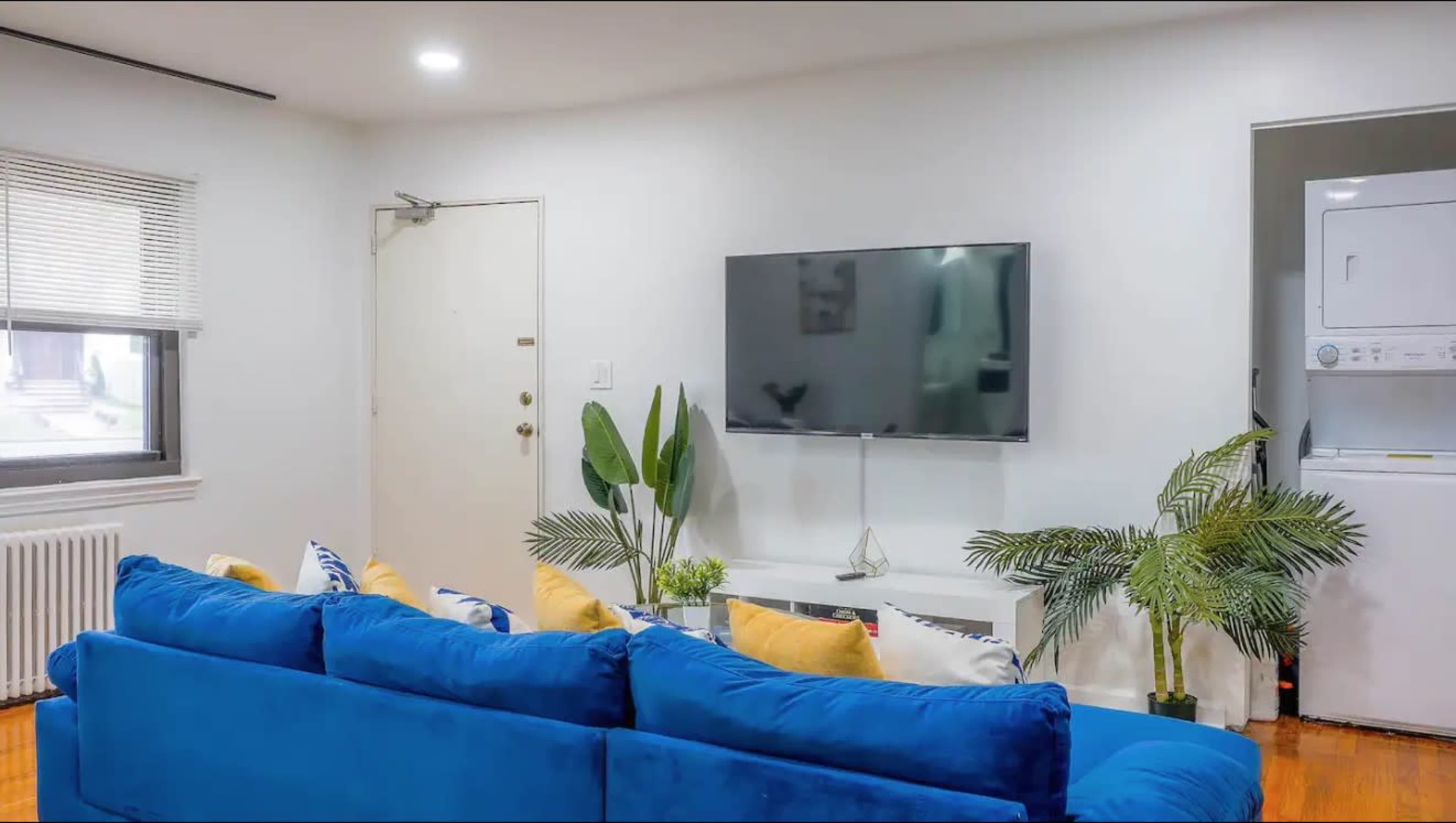 A living room features a blue sofa with yellow cushions, a wall-mounted television, and a plant beside a laundry area.