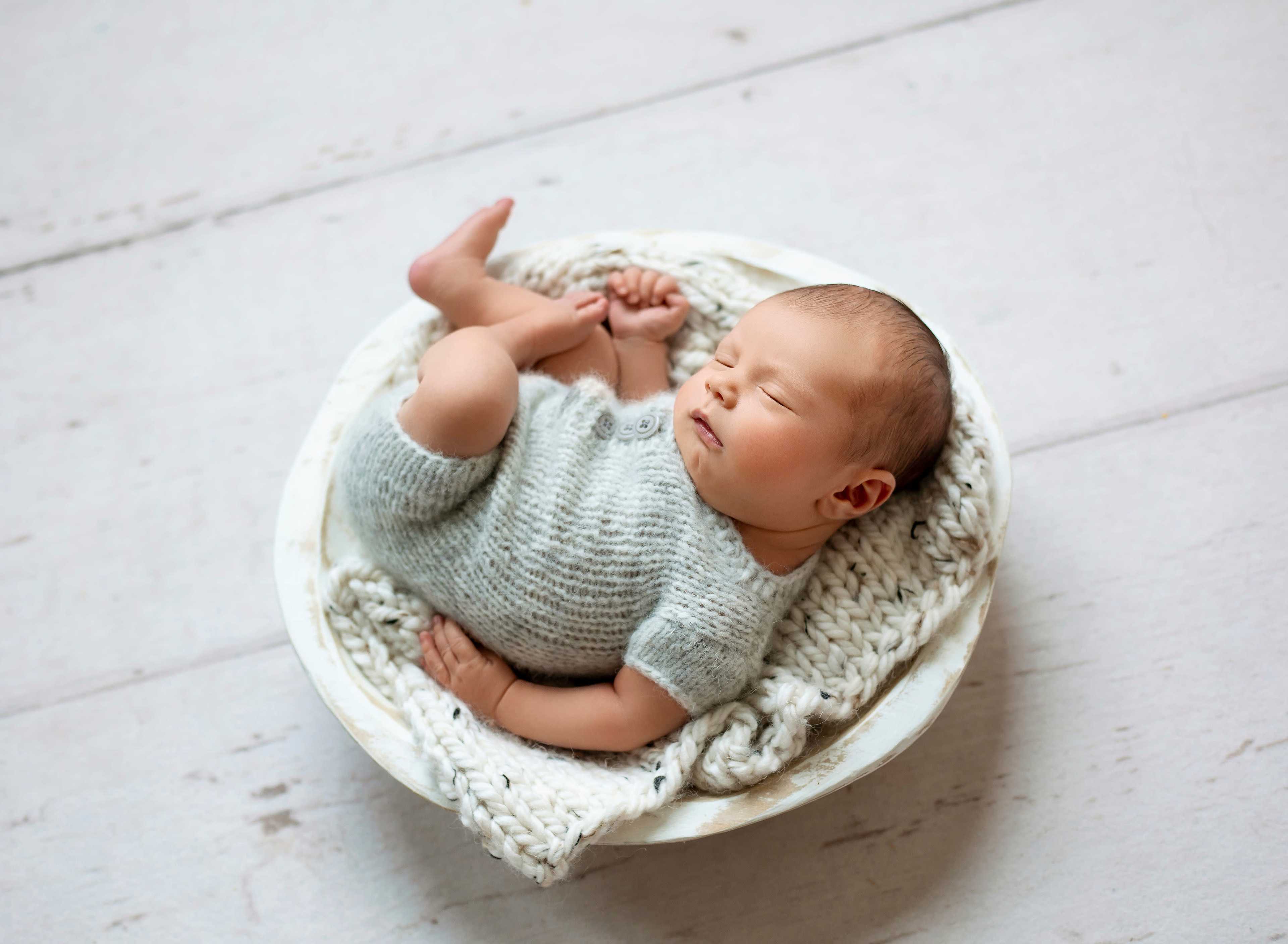 A sleeping baby is nestled in a round bowl lined with a knitted blanket.