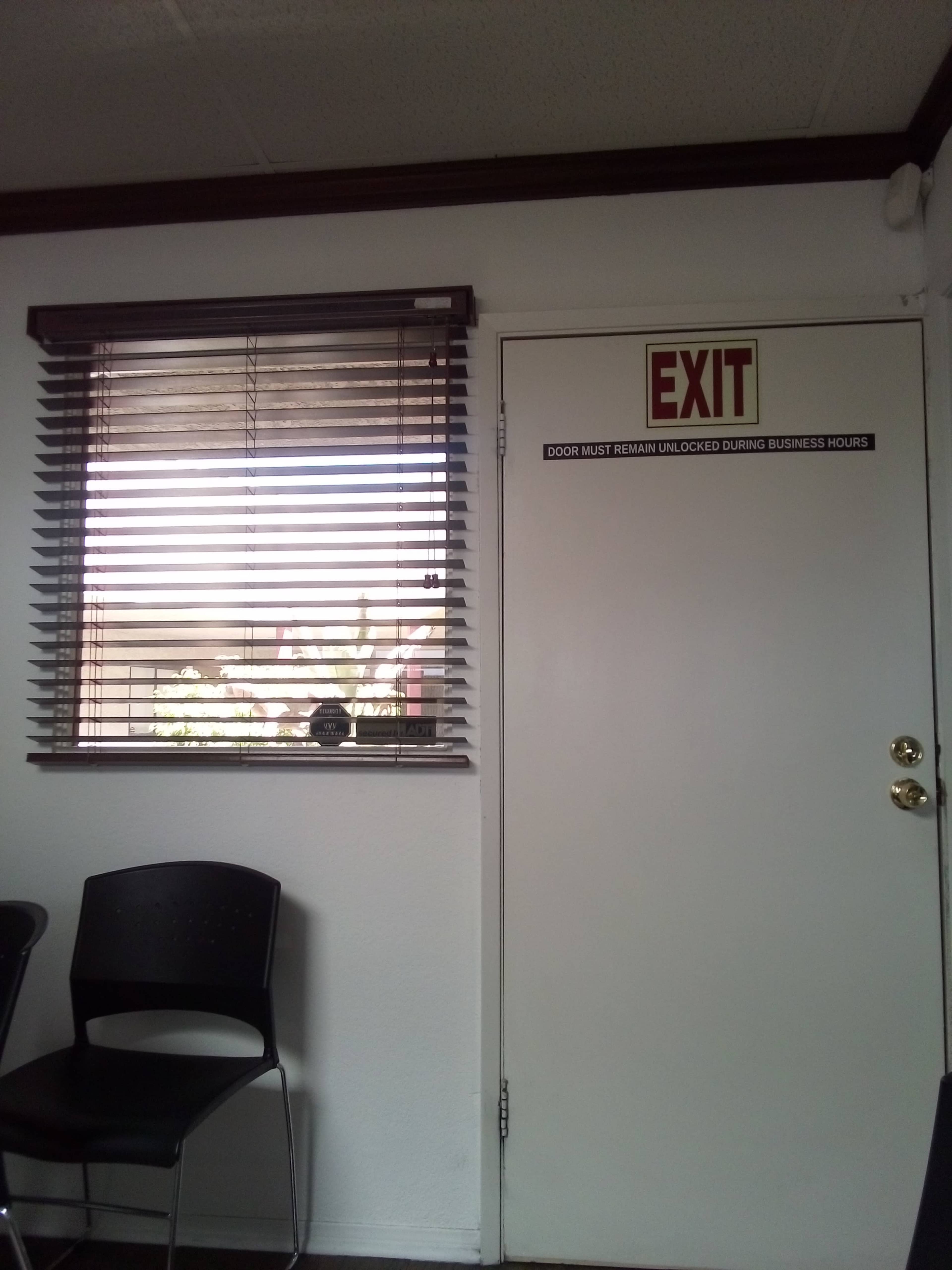 A closed exit door with a sign indicating it must remain unlocked during business hours is adjacent to a window covered with blinds.