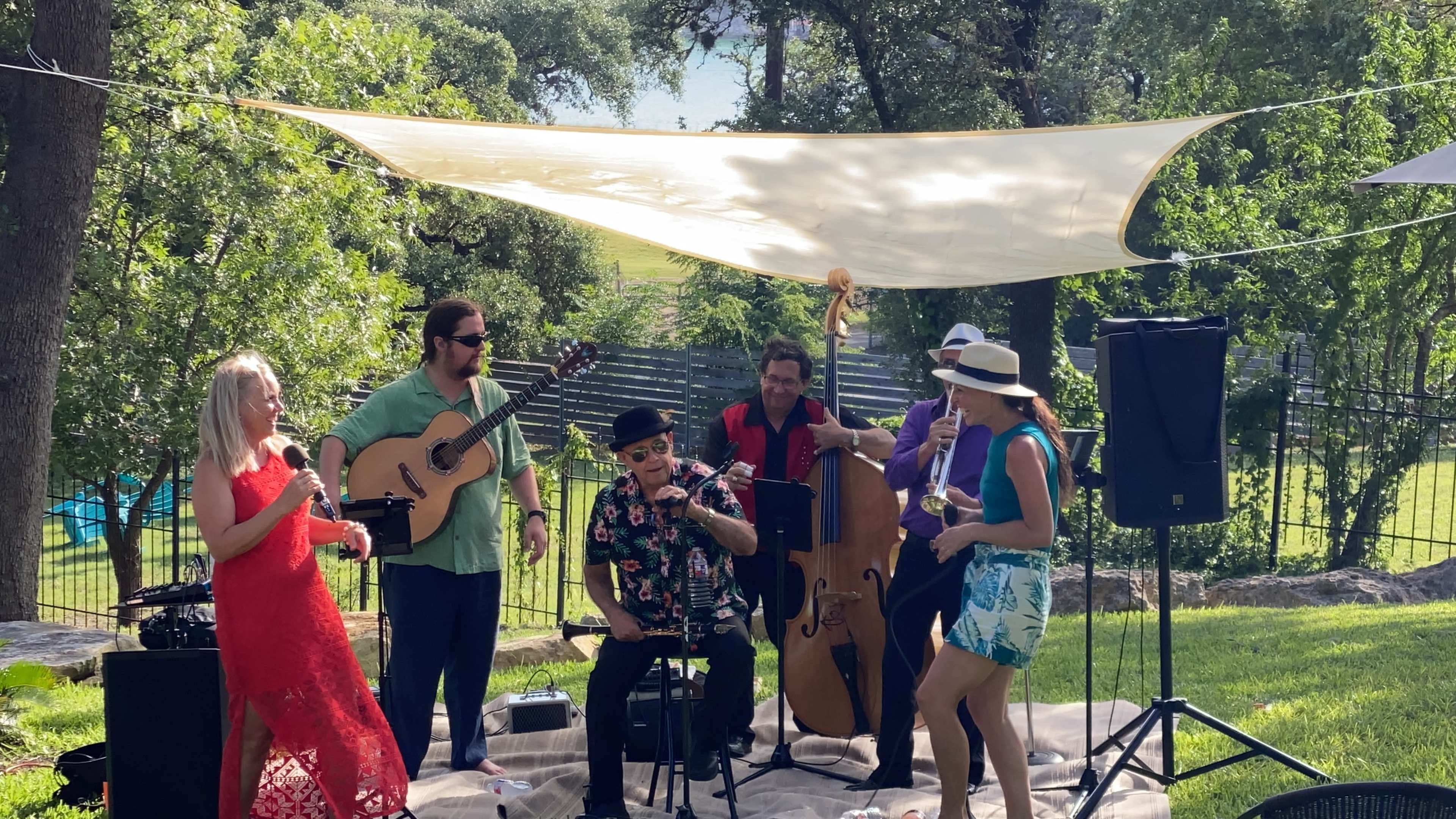 A live band performs on a grassy area under a fabric canopy, with musicians playing various instruments and a couple of performers singing.