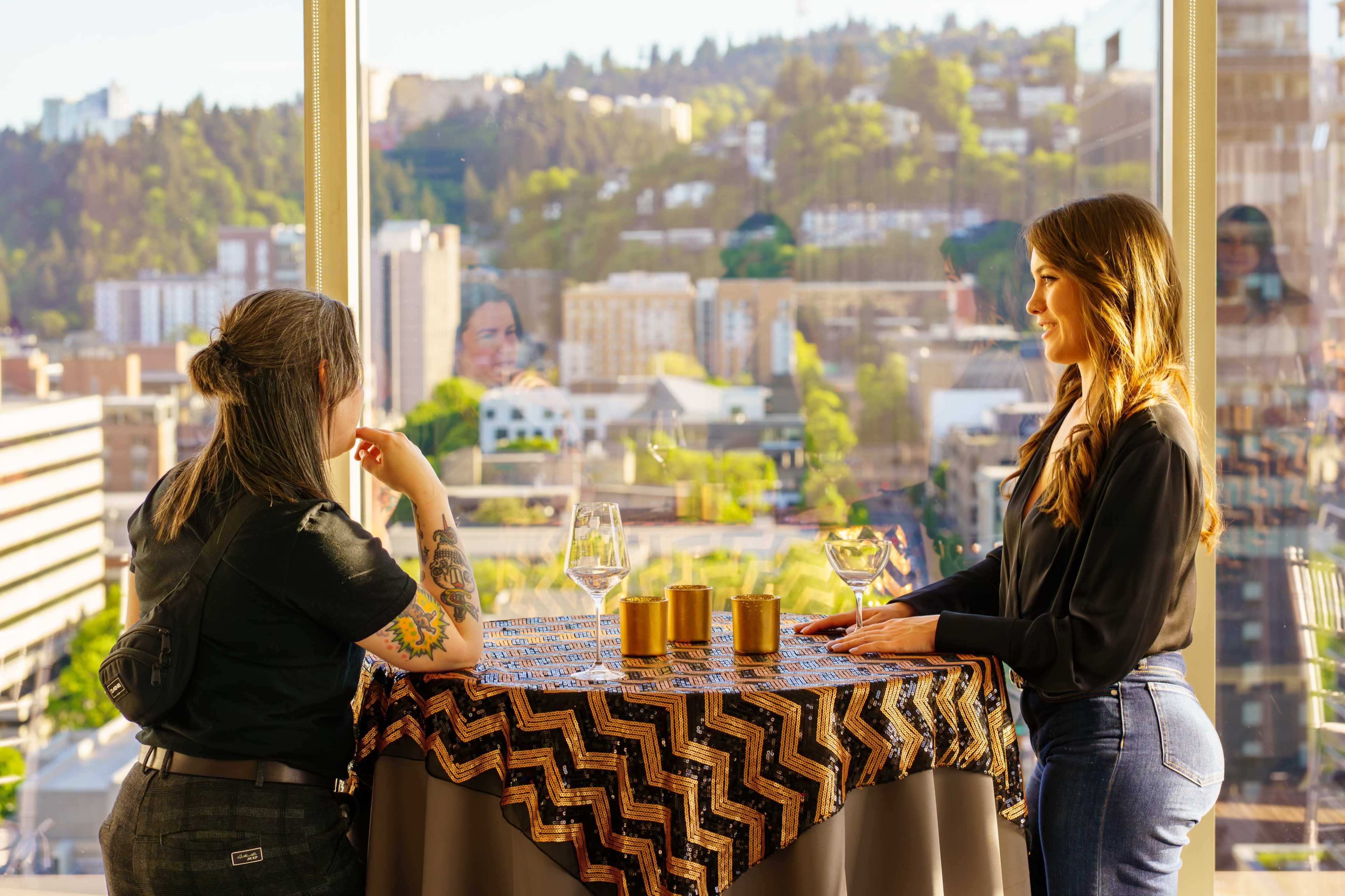 Two women are standing near a table with a decorative tablecloth and glasses, gazing out at a cityscape through a large window.