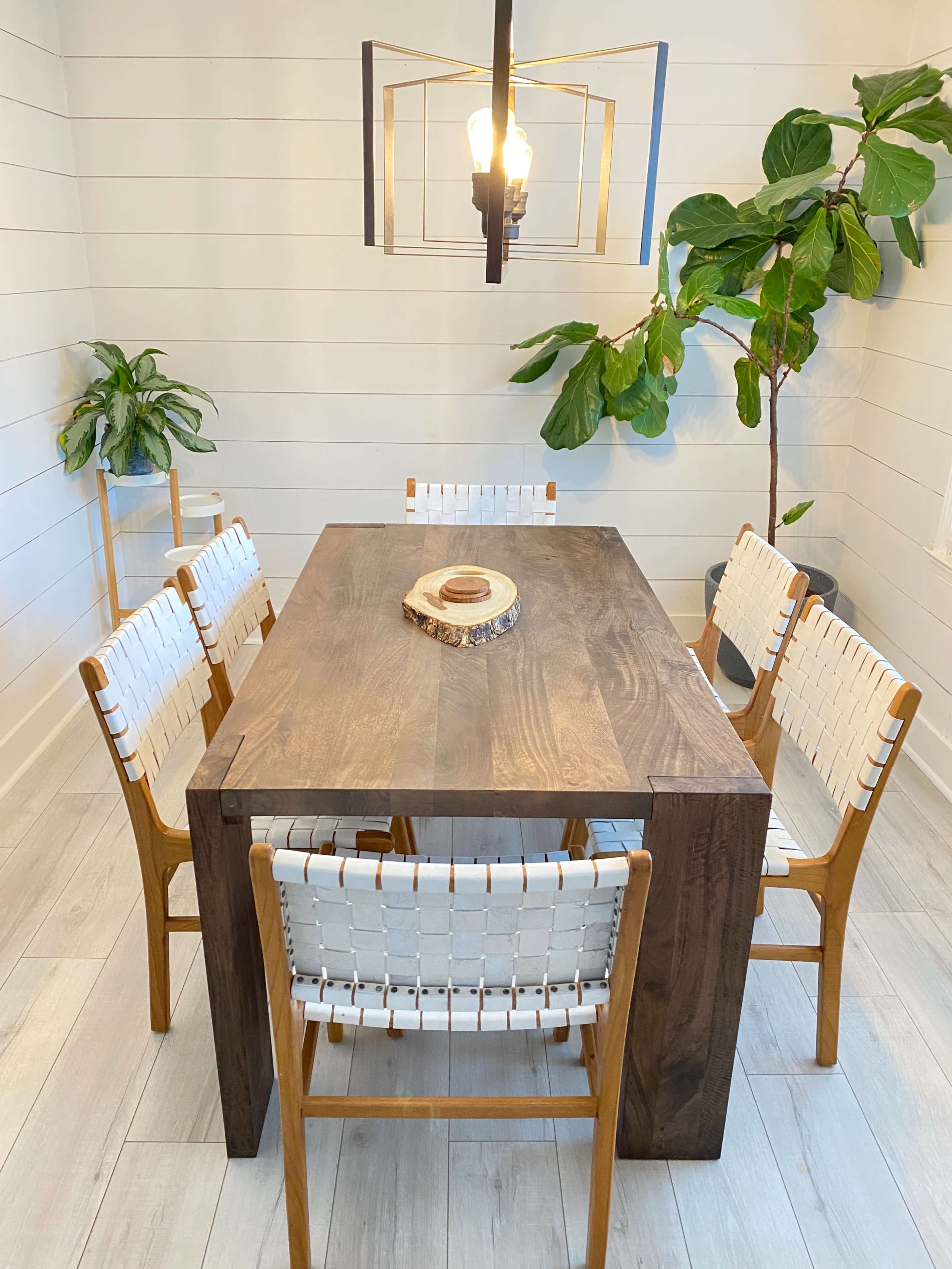 A wooden dining table is surrounded by six woven chairs in a bright, minimalist room with greenery.