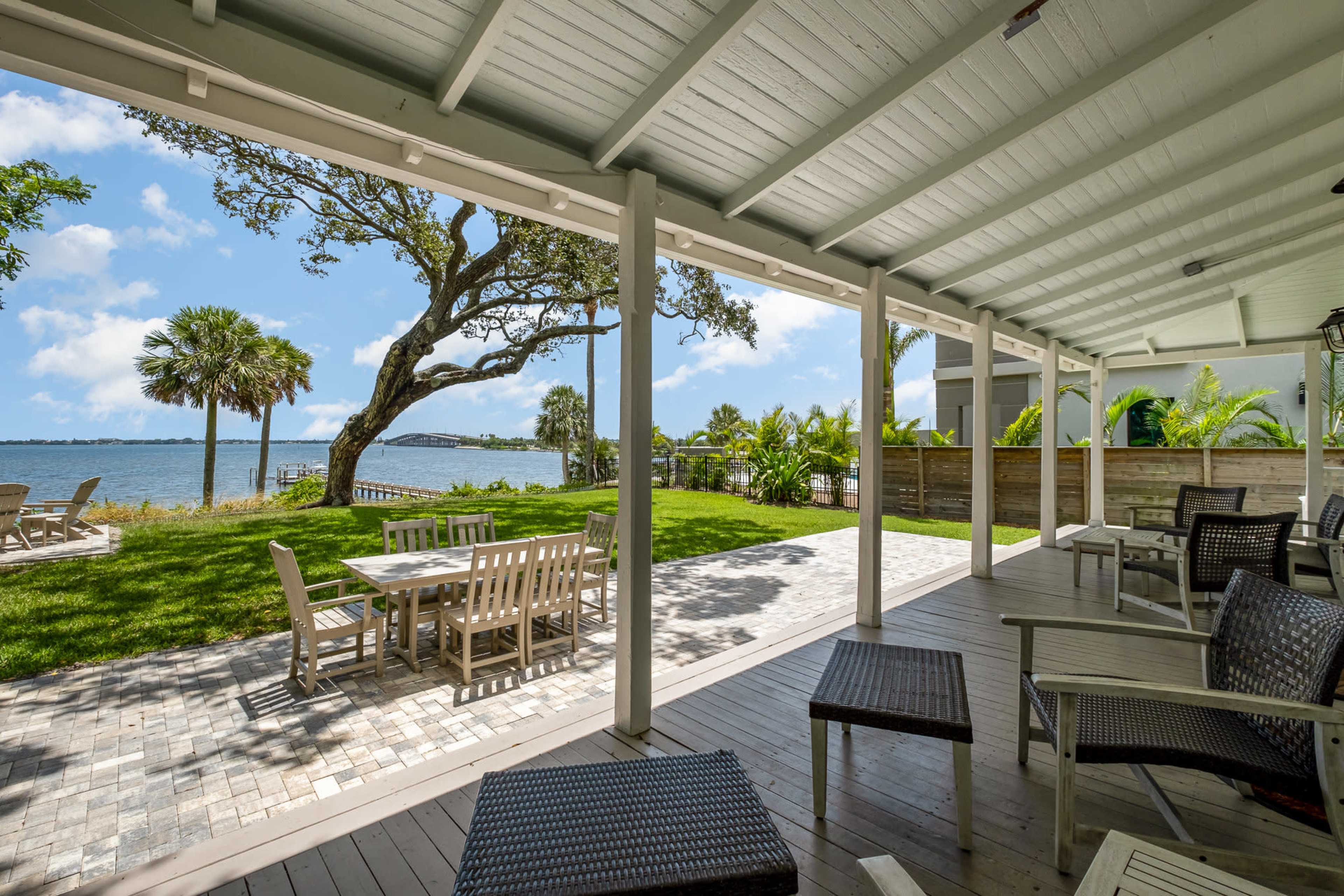 A shaded patio area with wooden furniture overlooks a grassy yard and a waterfront scene with palm trees.