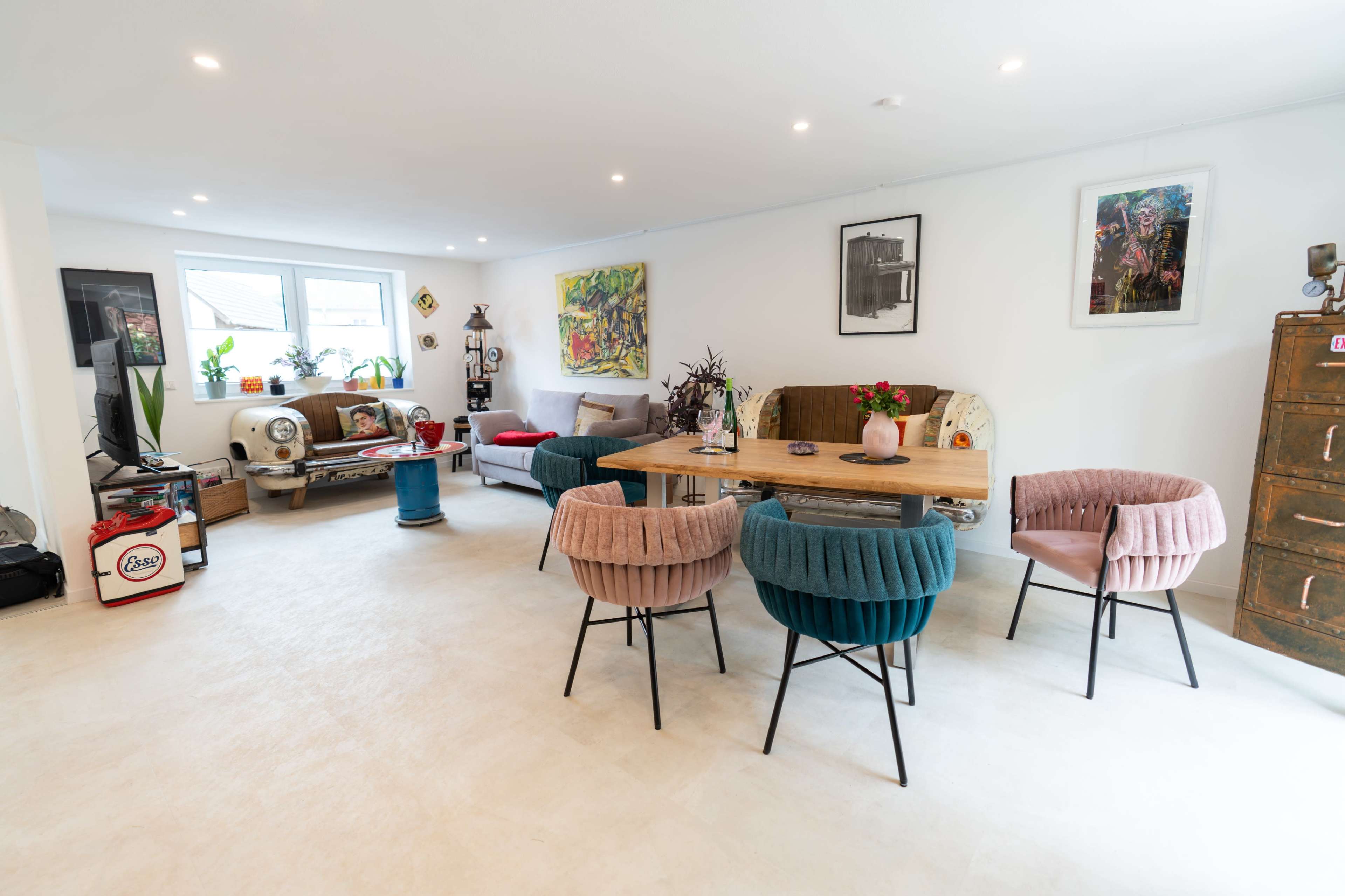 The image shows a spacious living area with a dining table surrounded by colorful chairs, art on the walls, and various decorative items scattered throughout the room.