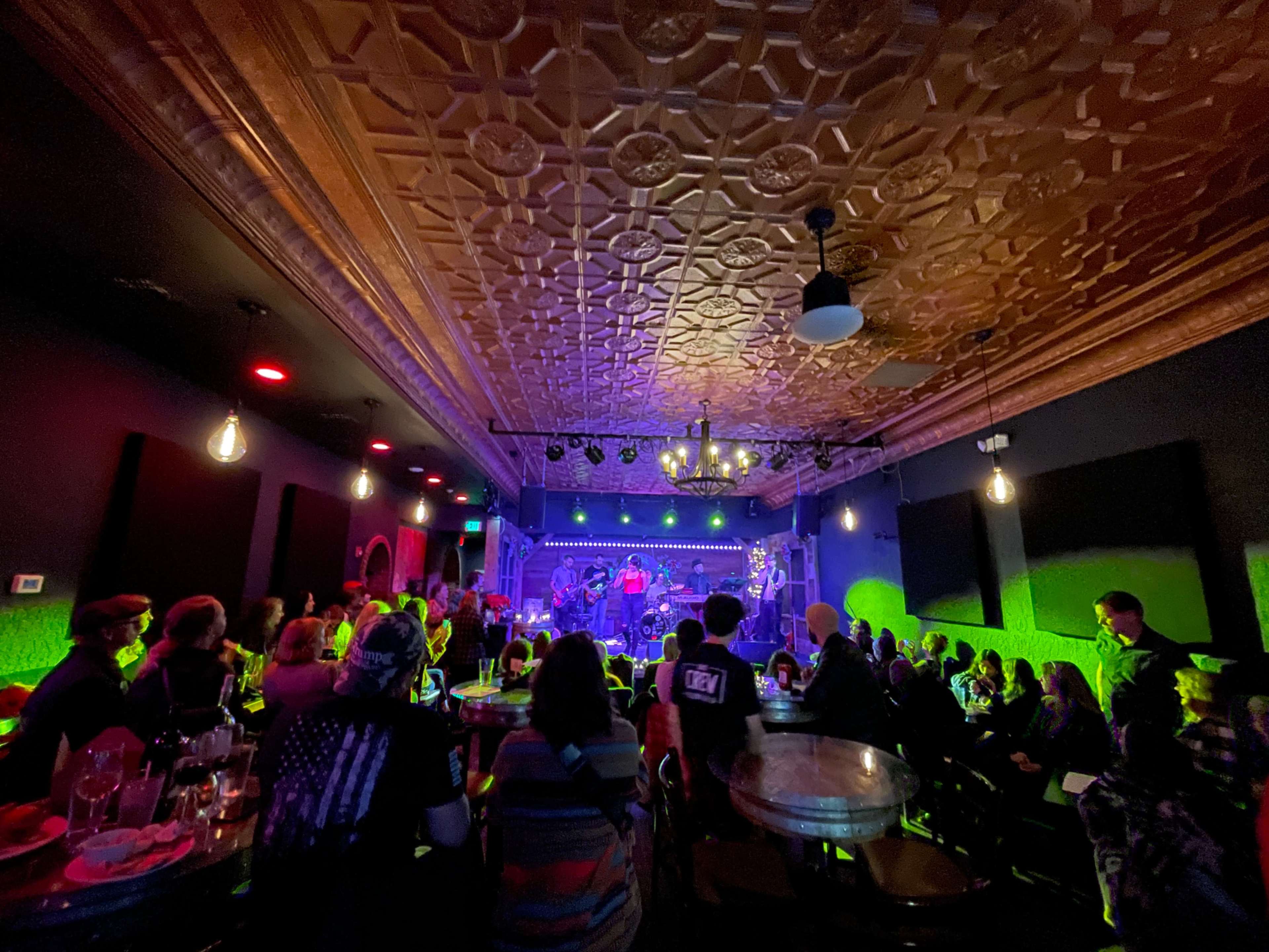 A crowded venue features a band performing on stage under colorful lighting, with patrons seated at tables.