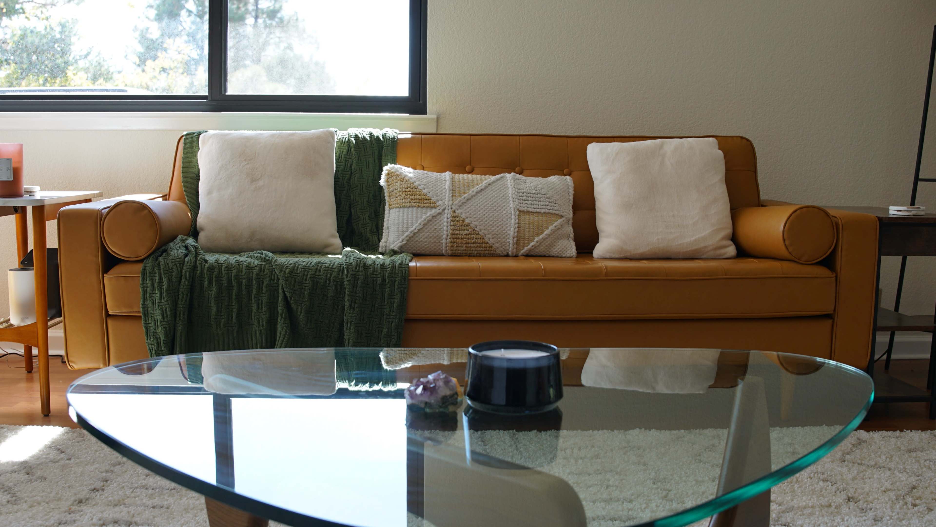 The scene features a brown leather sofa adorned with white and patterned pillows, accompanied by a green throw blanket, and is set in a room with a glass coffee table and a large window.