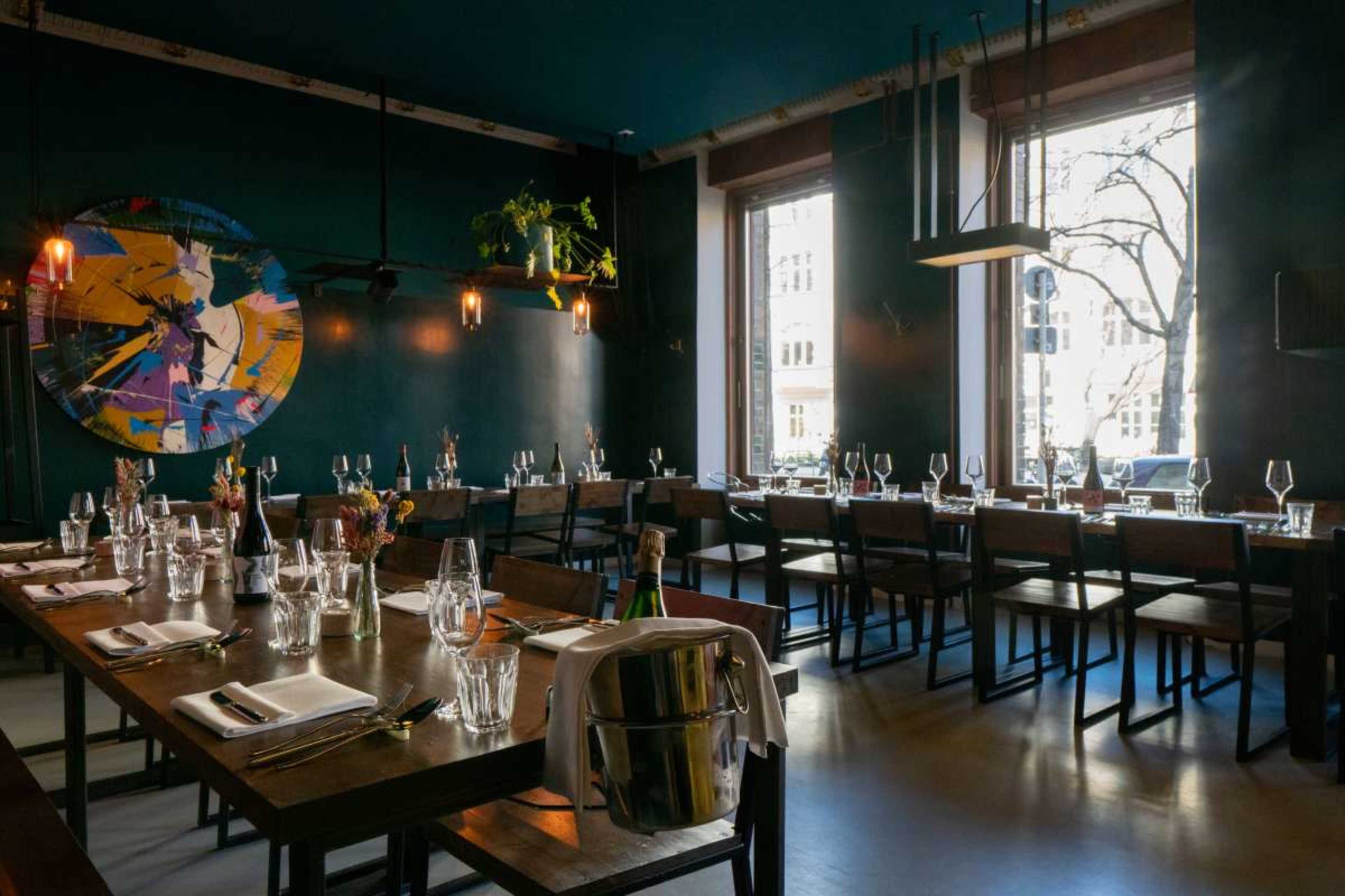 A dining area features long wooden tables set with glassware and tableware, and large windows providing natural light, along with a colorful abstract painting on the wall.