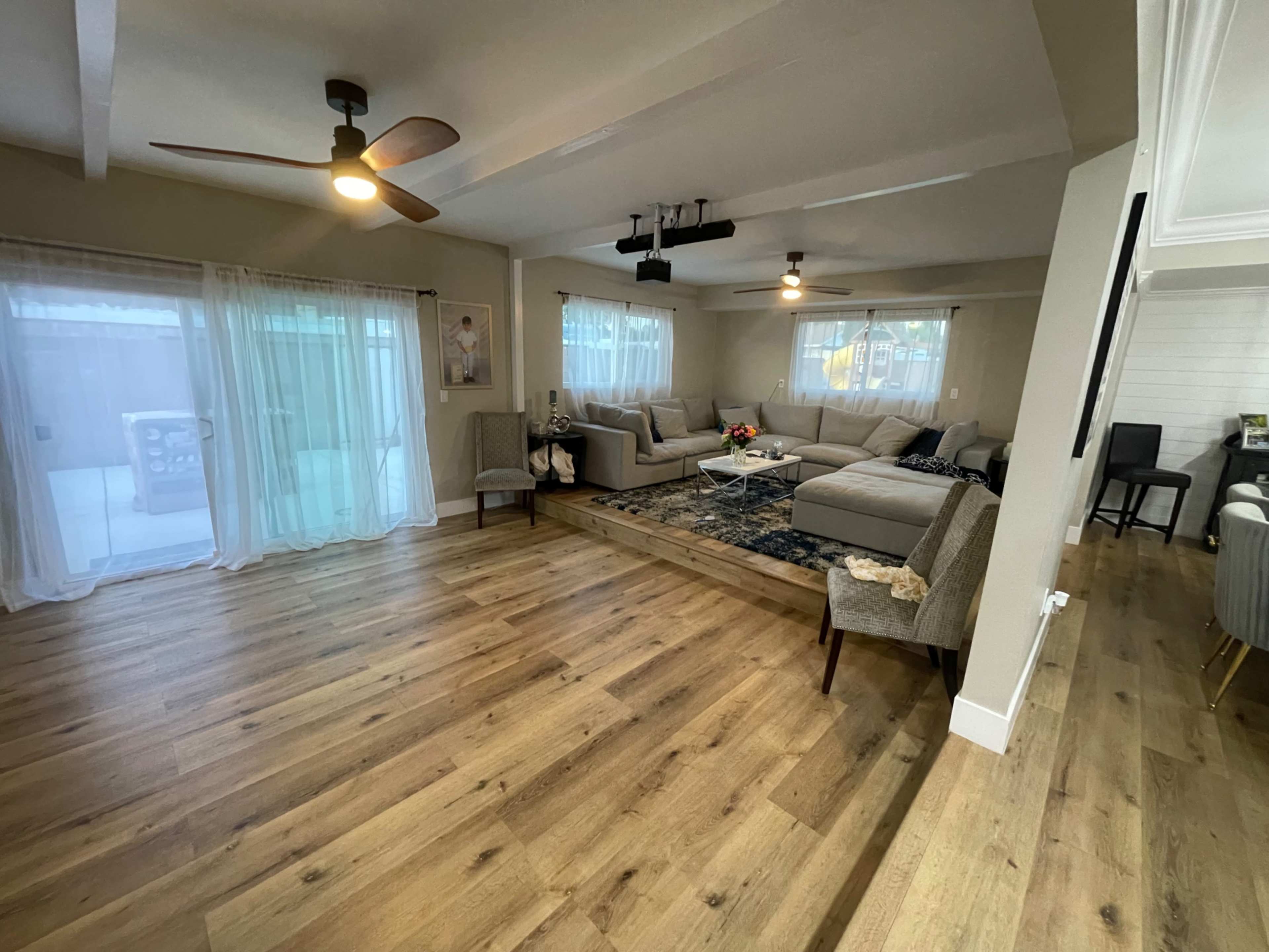 A spacious living room with a large sectional sofa, a coffee table centered on a patterned rug, and wooden flooring, illuminated by ceiling fans and natural light through large windows.