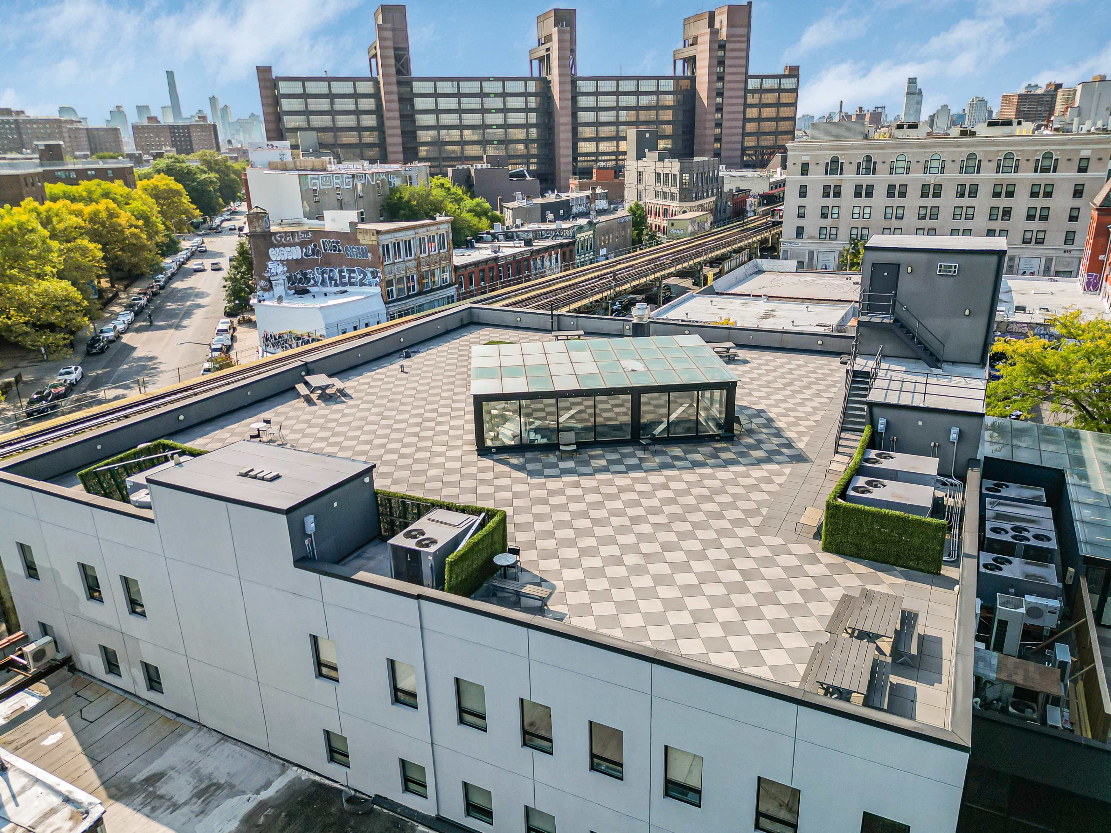The image shows an aerial view of a large, flat rooftop in an urban area featuring a glass-enclosed structure, greenery, and mechanical units, with a backdrop of city buildings and an elevated train track.
