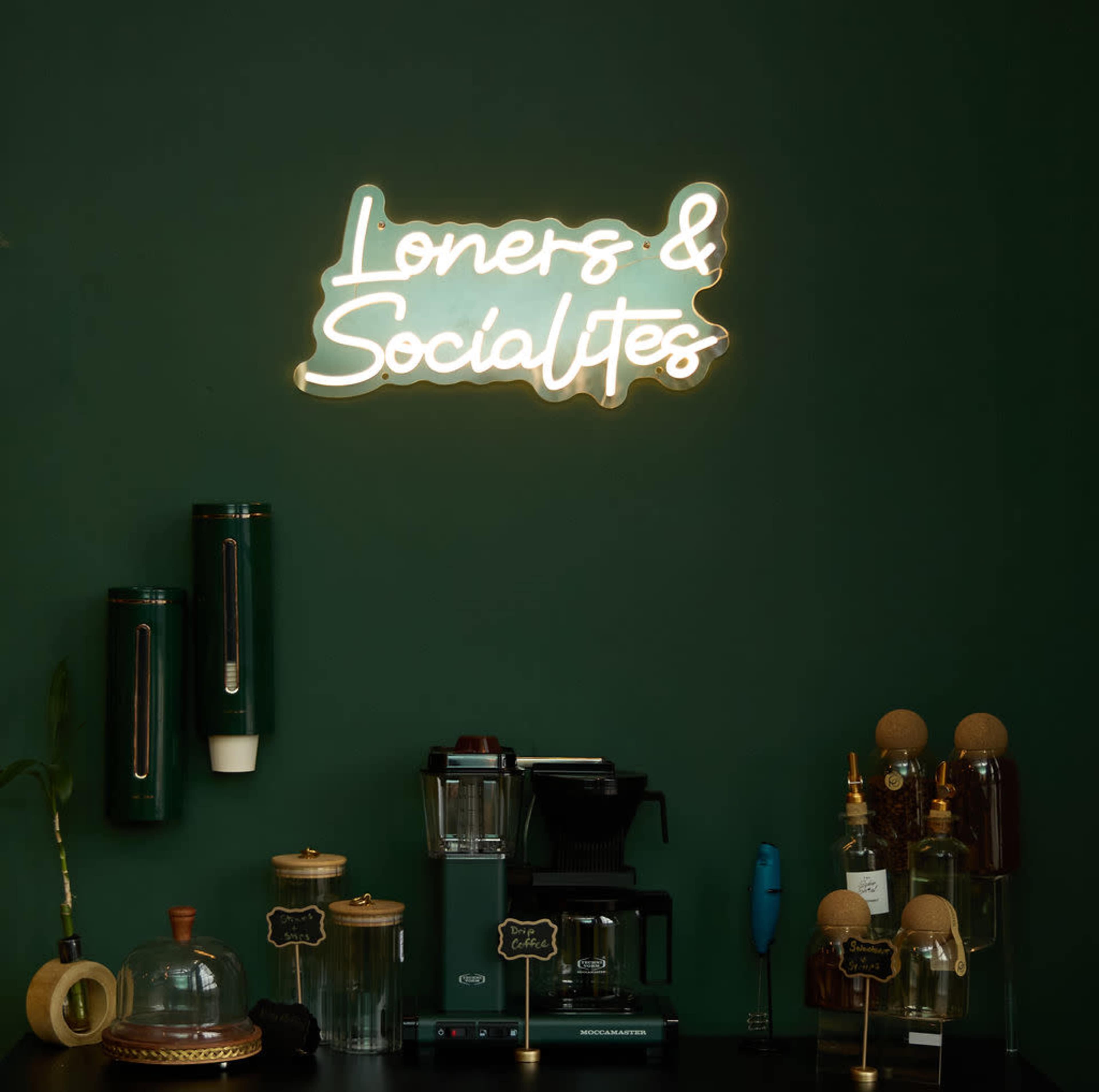 A dark green wall with a neon sign that reads "Loners & Socialites" above a coffee-making setup and various kitchen utensils.