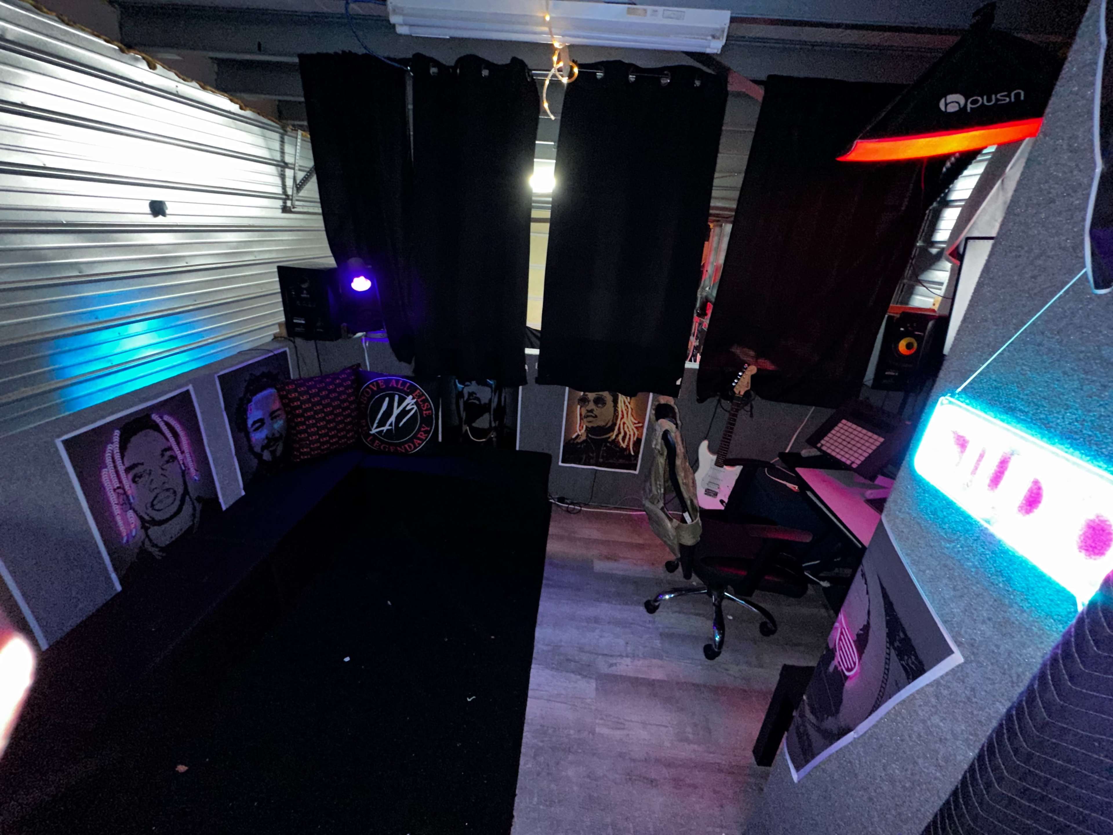 A small, dimly lit room with black curtains, a desk with a computer and chair, and several framed artworks on the walls.