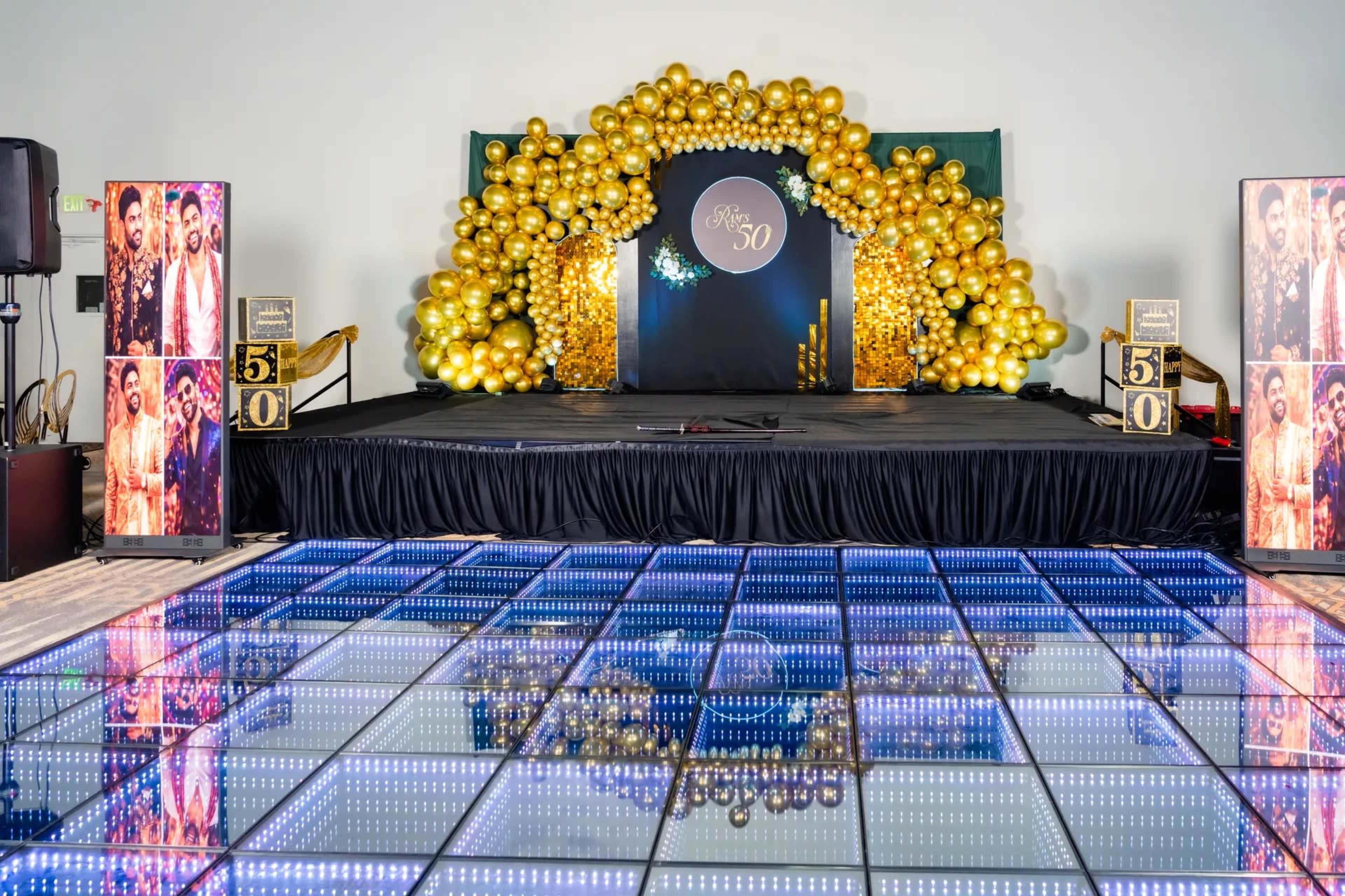 The scene features a stage decorated with a backdrop of golden balloons, illuminated by a light-up dance floor and adorned with framed portraits of men on either side.