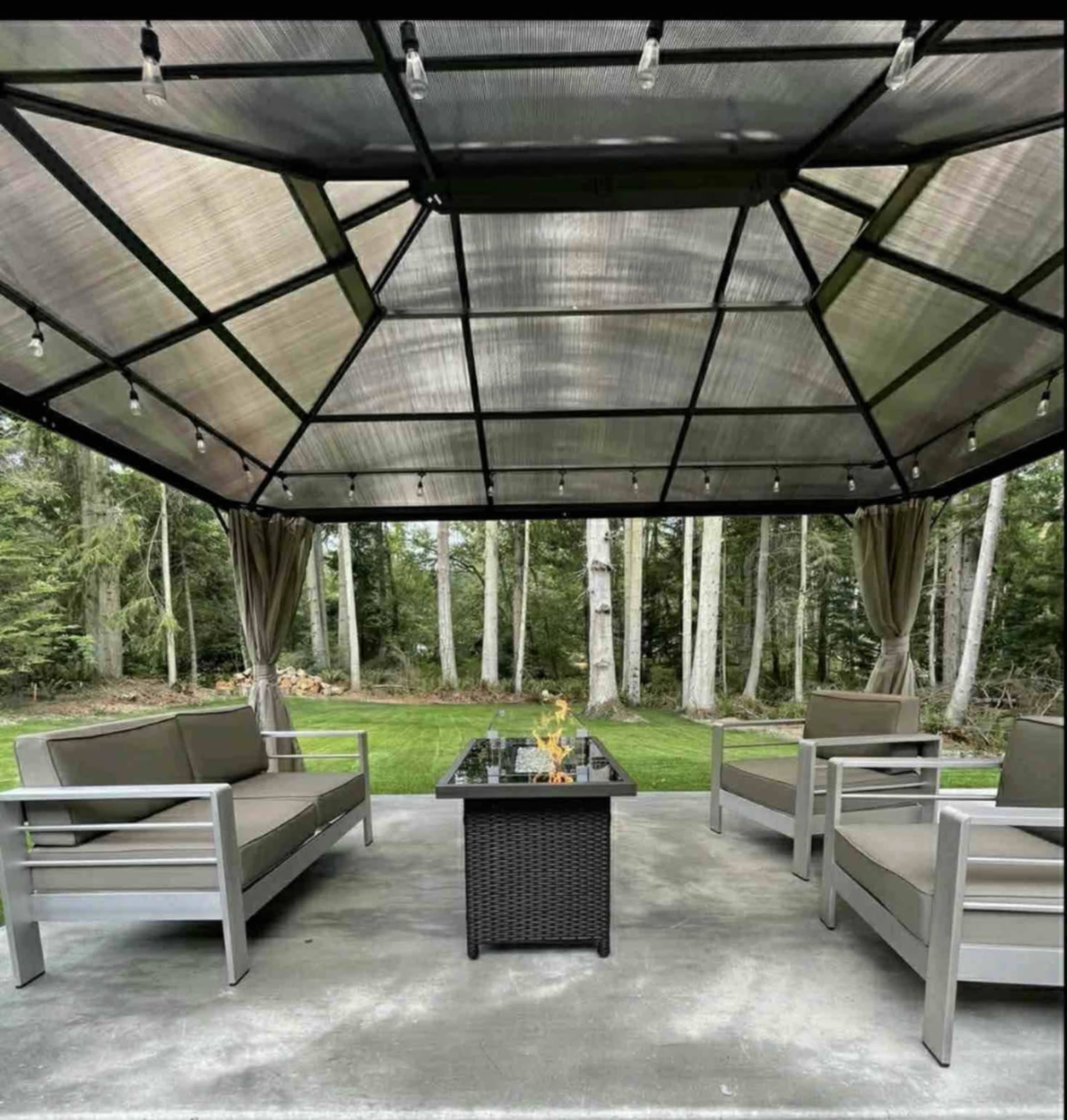 A covered outdoor seating area with four gray couches arranged around a central fire table surrounded by trees.