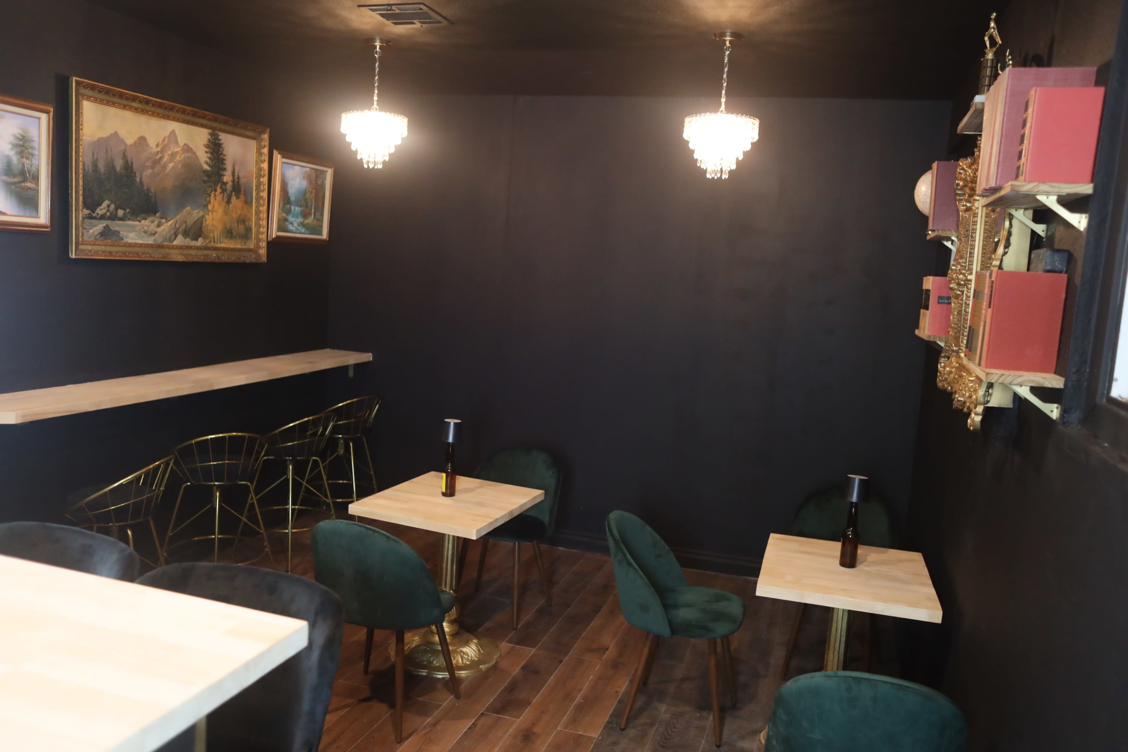 The image shows a dimly lit café with dark walls, wooden flooring, and small tables surrounded by green chairs, along with a bar area and decorative wall pieces.