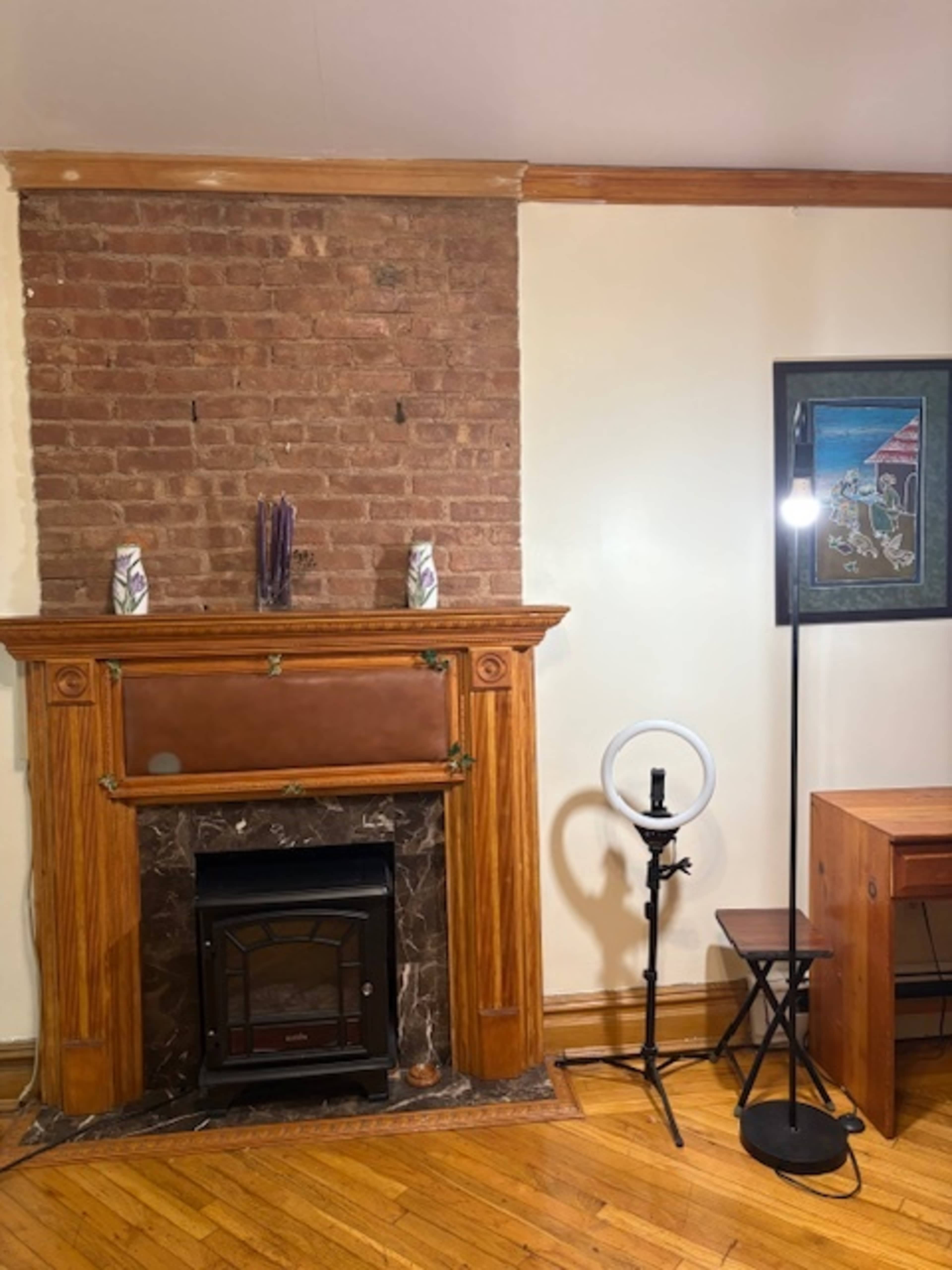 The image shows a room with a brick wall, a wooden fireplace with decorative elements, a ring light on a stand, and a small wooden table.