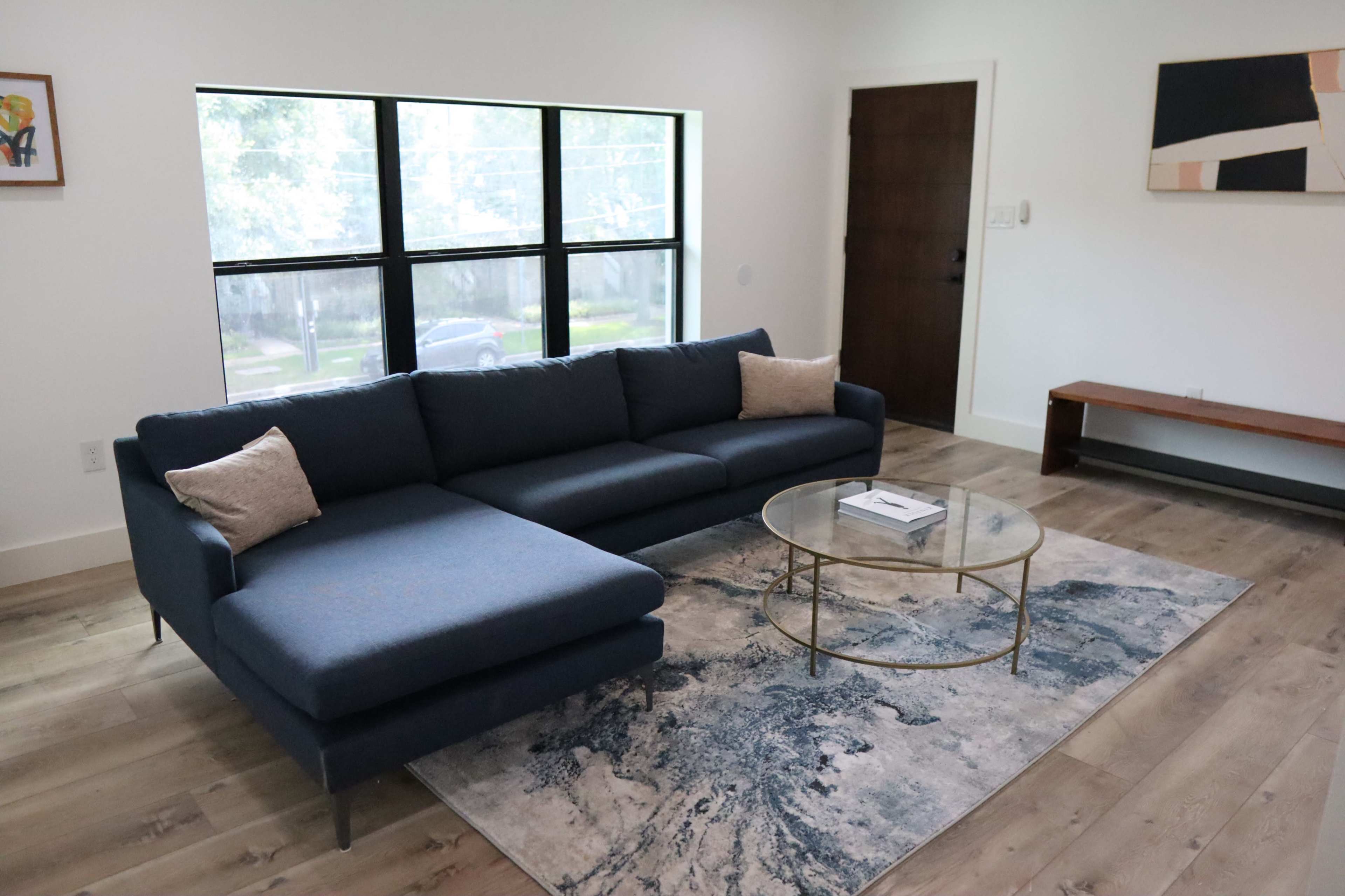 A modern living room featuring a blue sectional sofa, a round glass coffee table, and a rug on hardwood flooring, illuminated by natural light through large windows.