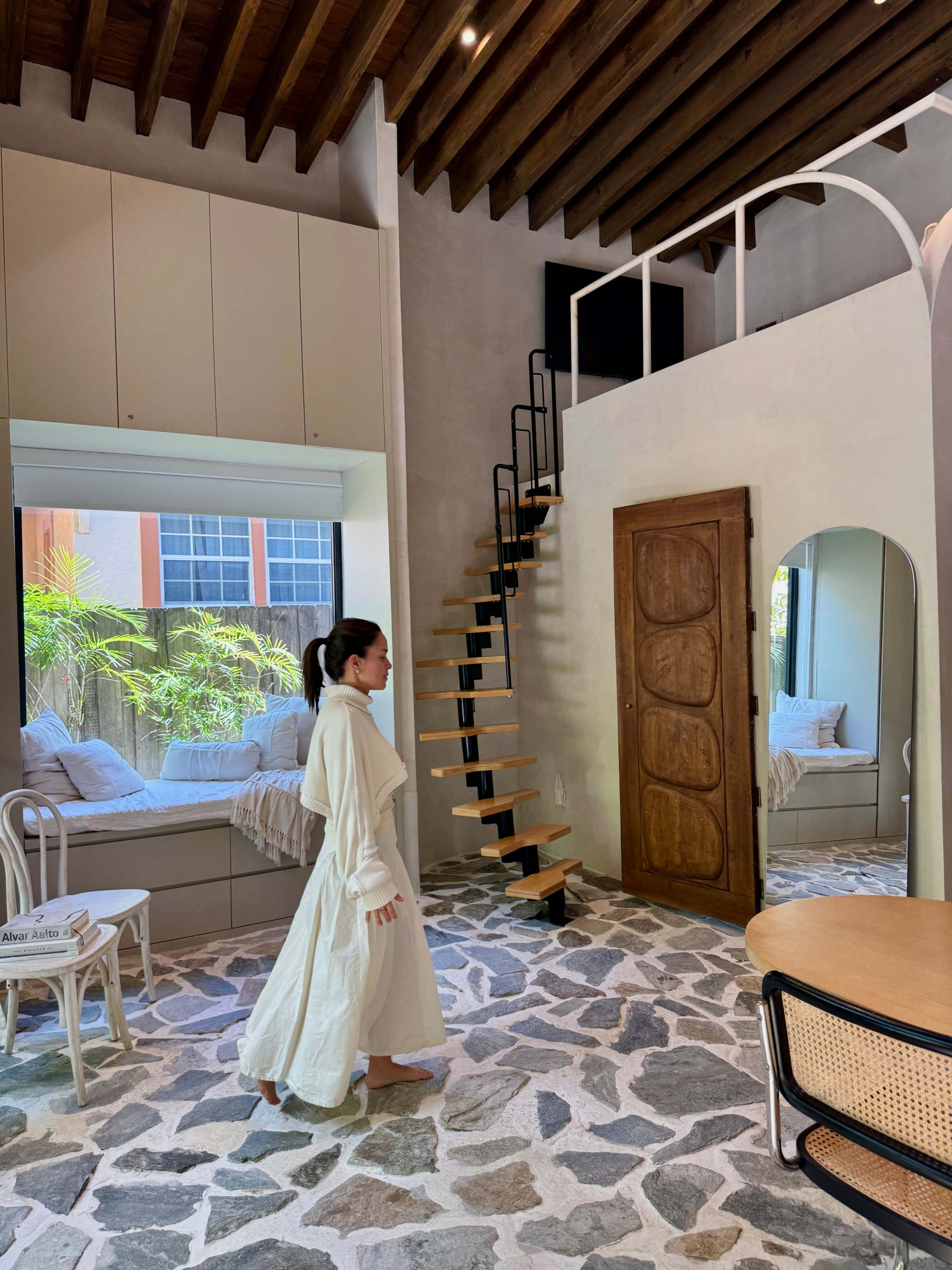 A woman in a white outfit walks past a wooden staircase in a modern living space featuring stone flooring and large windows.