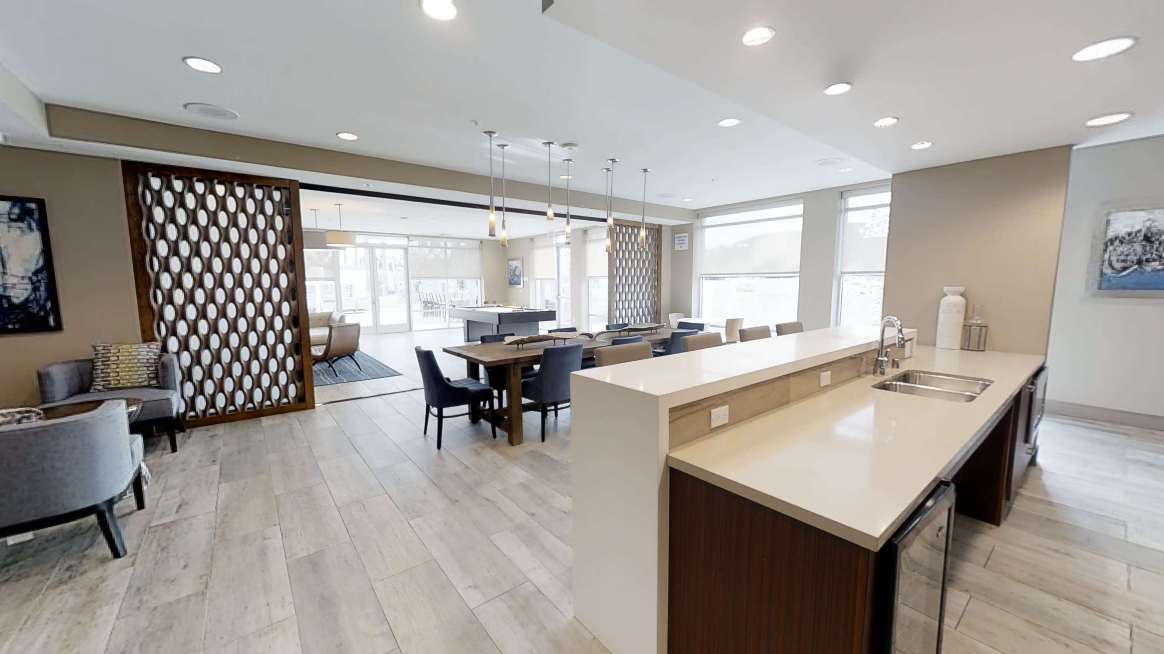 A modern, open-concept communal space with a kitchen area and seating arrangements, featuring large windows that allow natural light to fill the room.