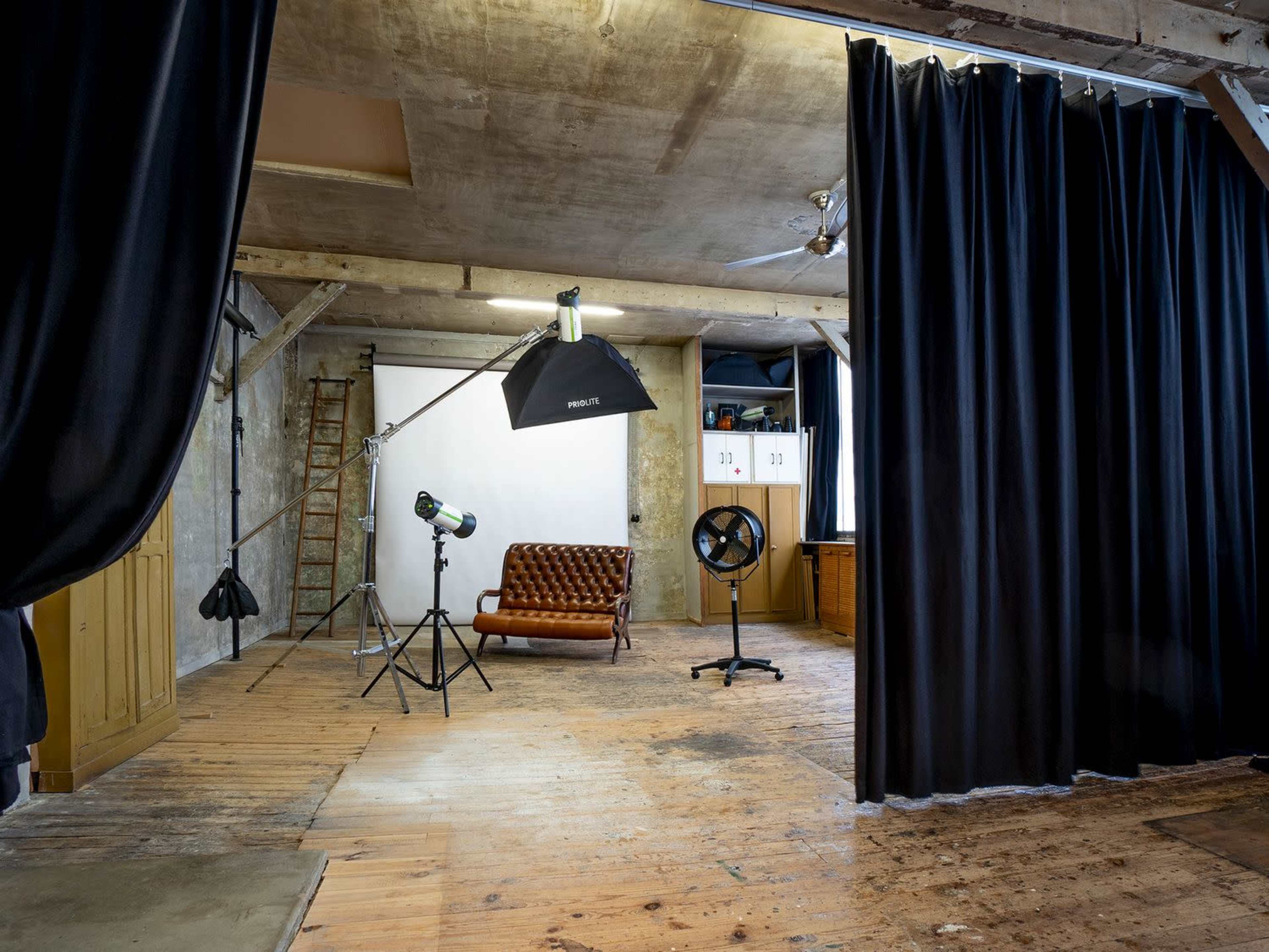 A photography studio features a brown leather sofa, lighting equipment, and black curtains in a spacious room with wooden floors and exposed concrete walls.