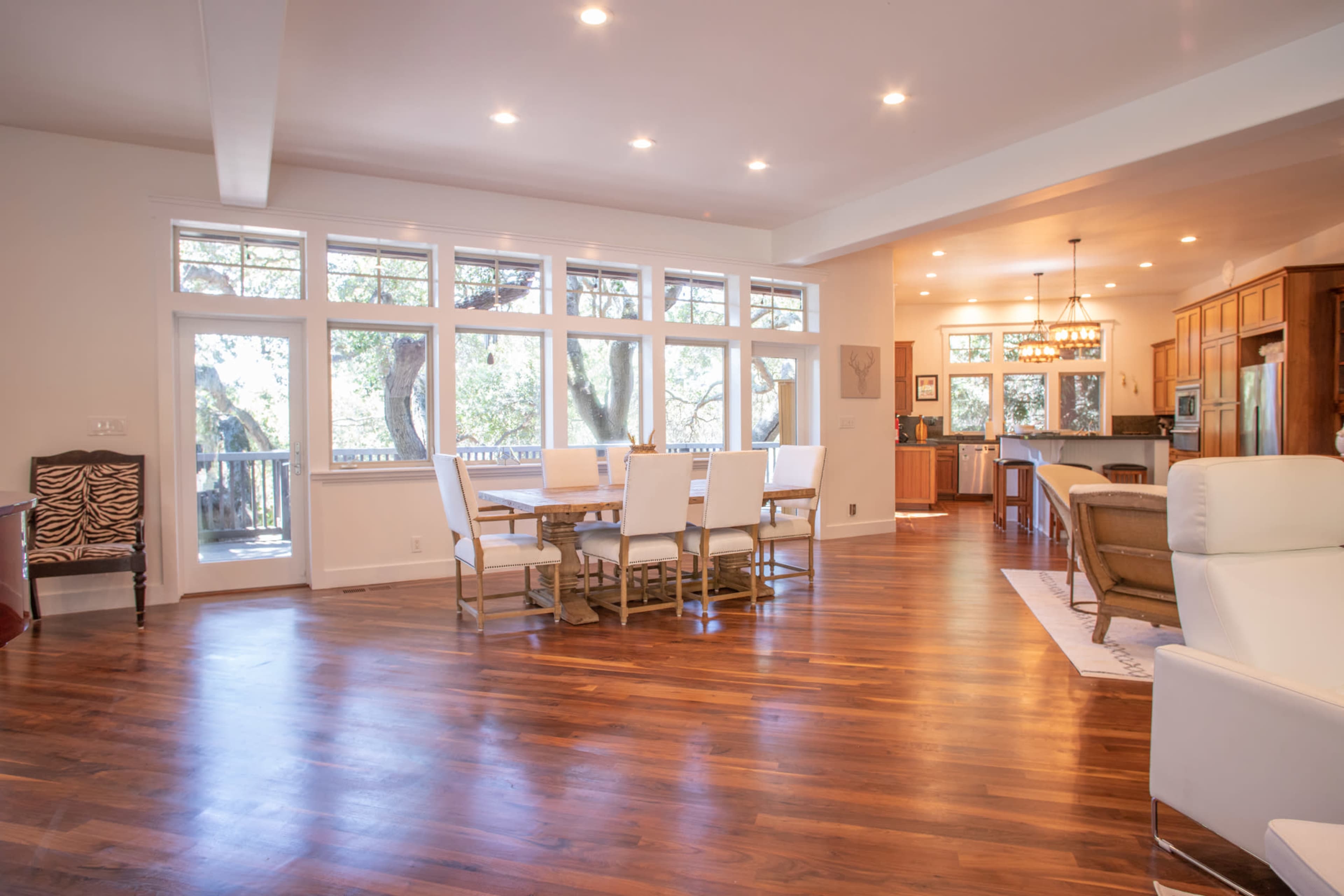 A spacious living area with hardwood floors, large windows, a dining table, and a kitchen in the background.