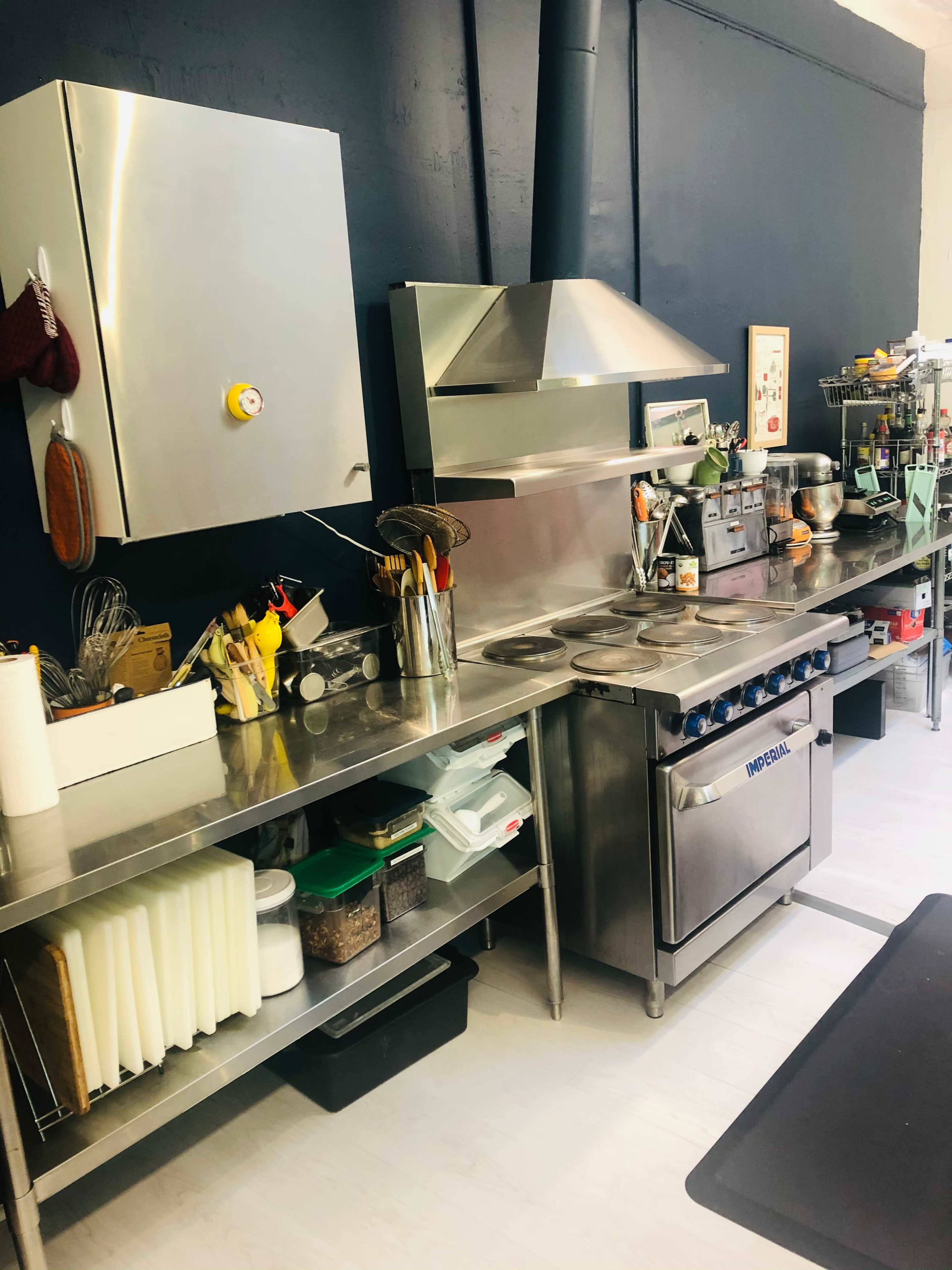 The image shows a stainless steel kitchen workspace with various cooking utensils, appliances, and storage containers against a dark wall.