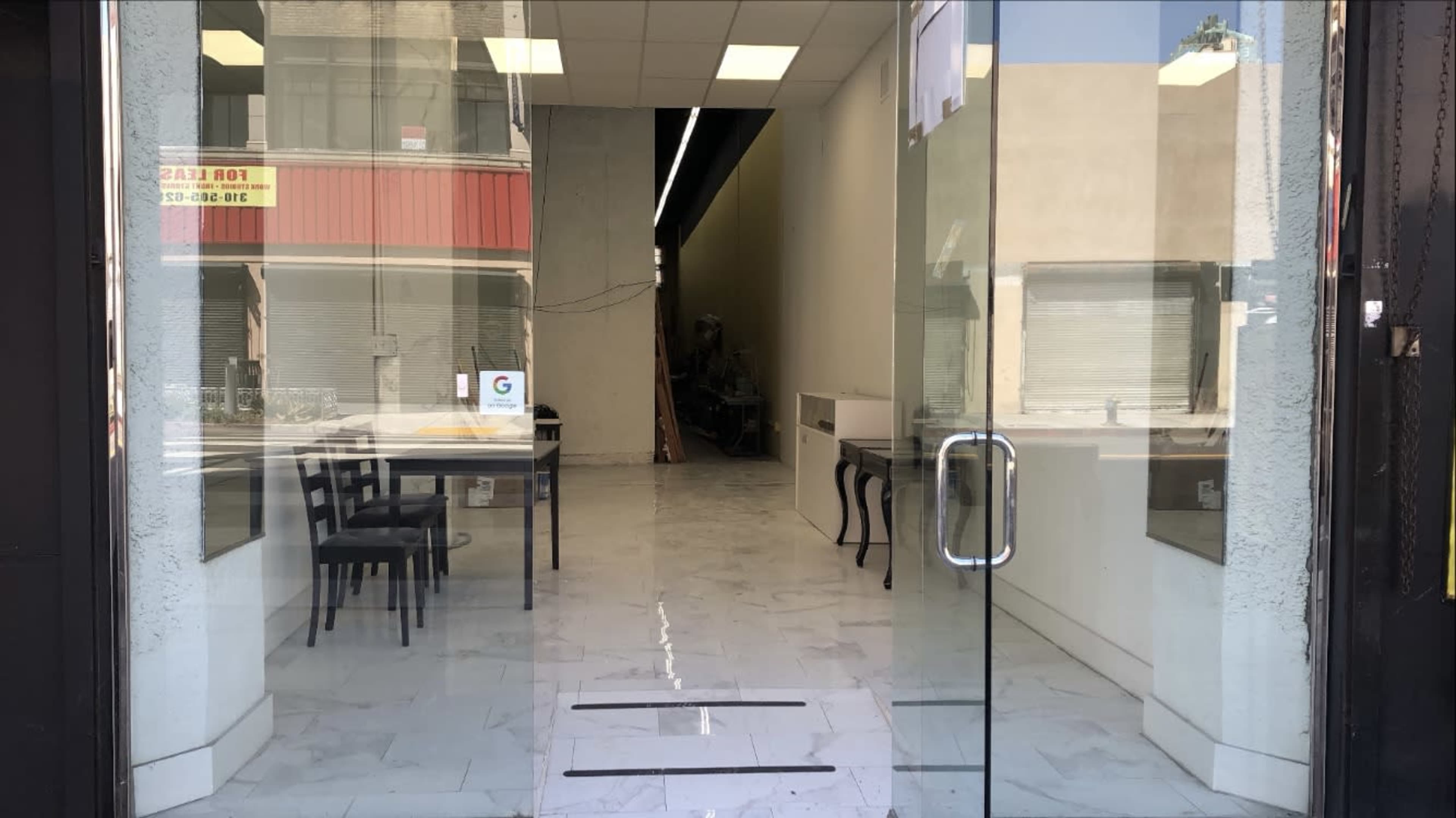The image shows the interior of a vacant storefront with a tiled floor and a few chairs visible through the glass doors.