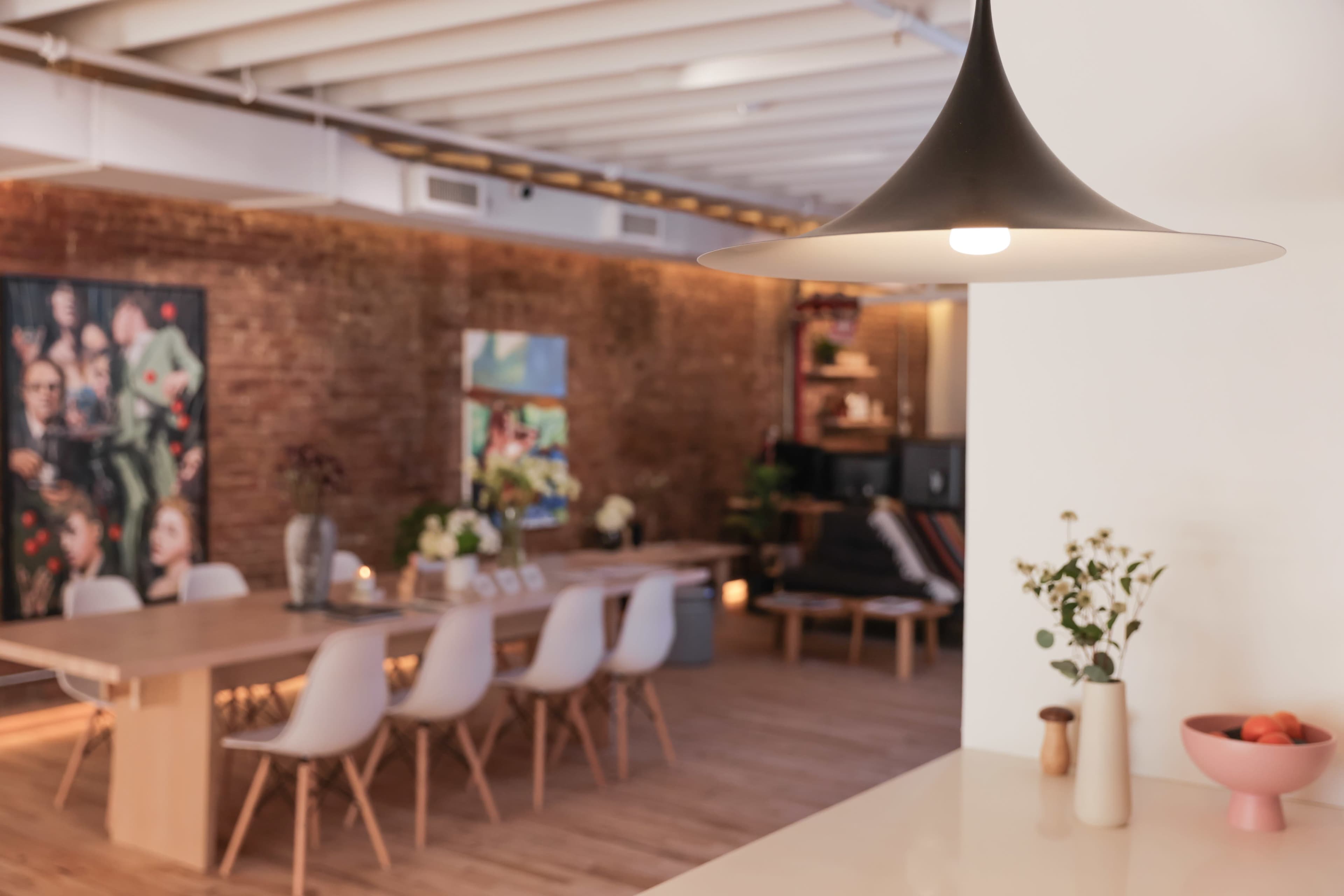 The image shows a modern interior with a dining table surrounded by chairs, decorative plants, and a pendant light hanging from the ceiling.
