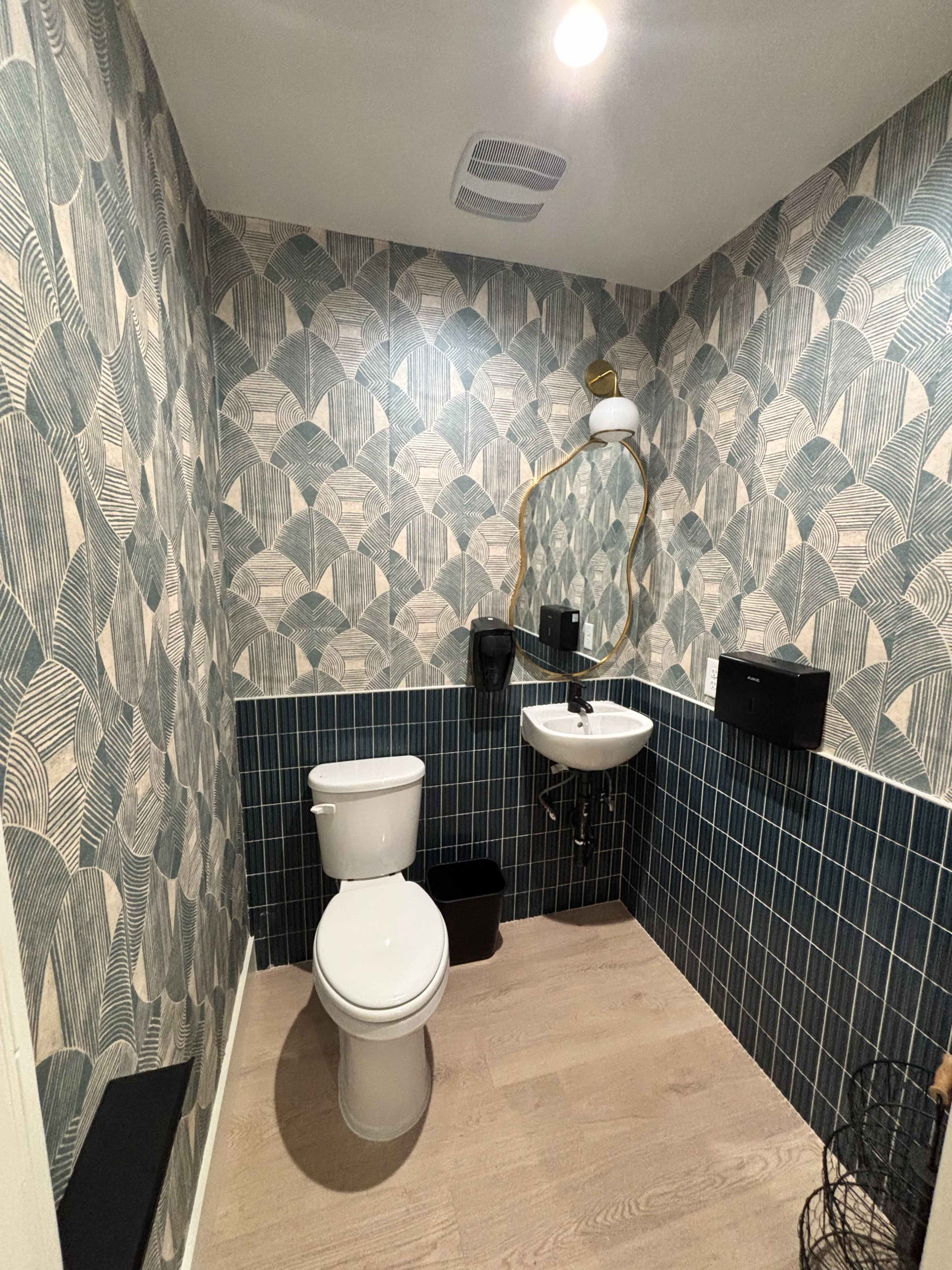 The image shows a small bathroom featuring patterned wallpaper, a toilet, a wall-mounted sink, and a modern light fixture.