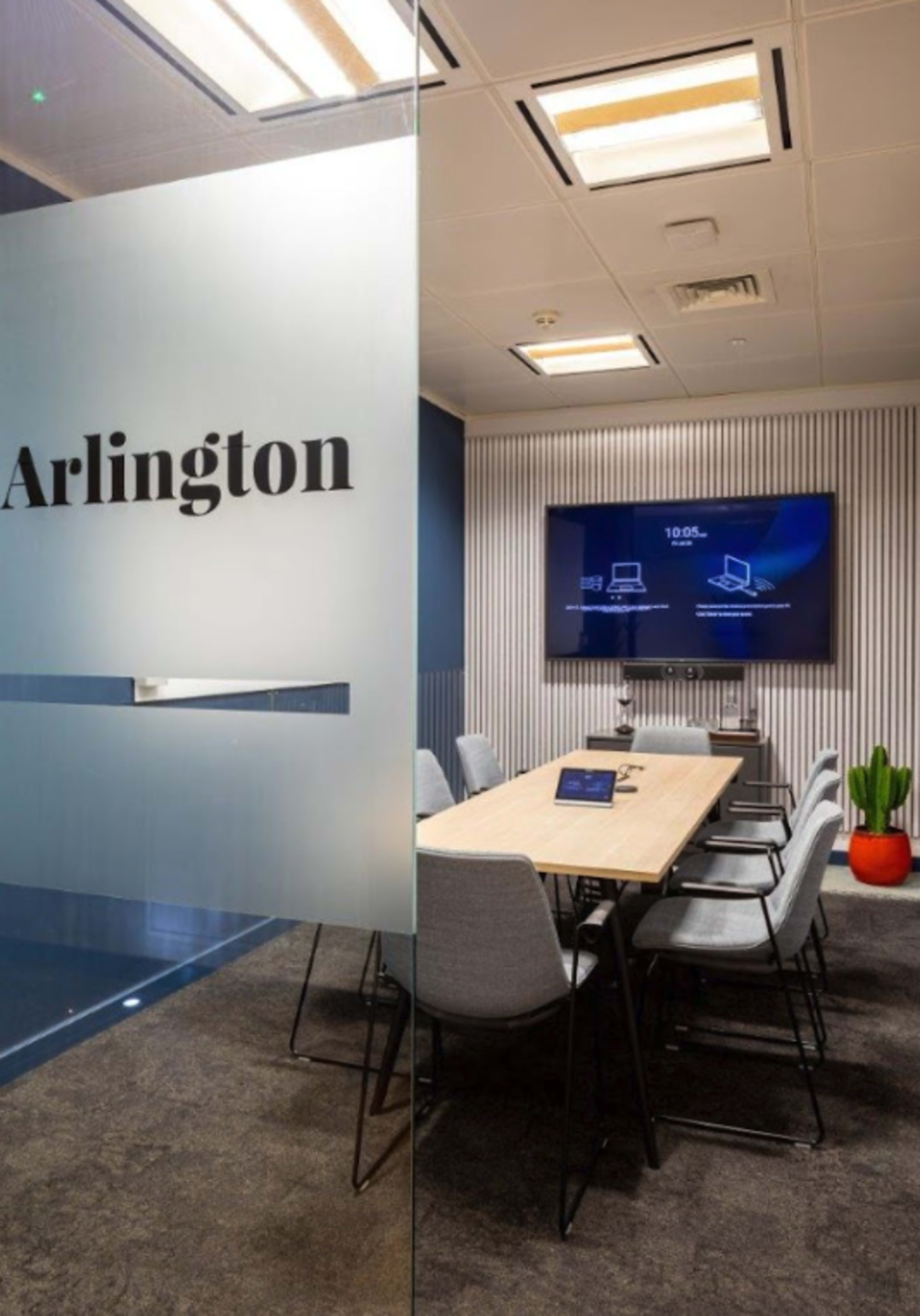 The image shows a modern conference room with a long wooden table, gray chairs, and a wall-mounted display screen, labeled "Arlington" on the glass door.