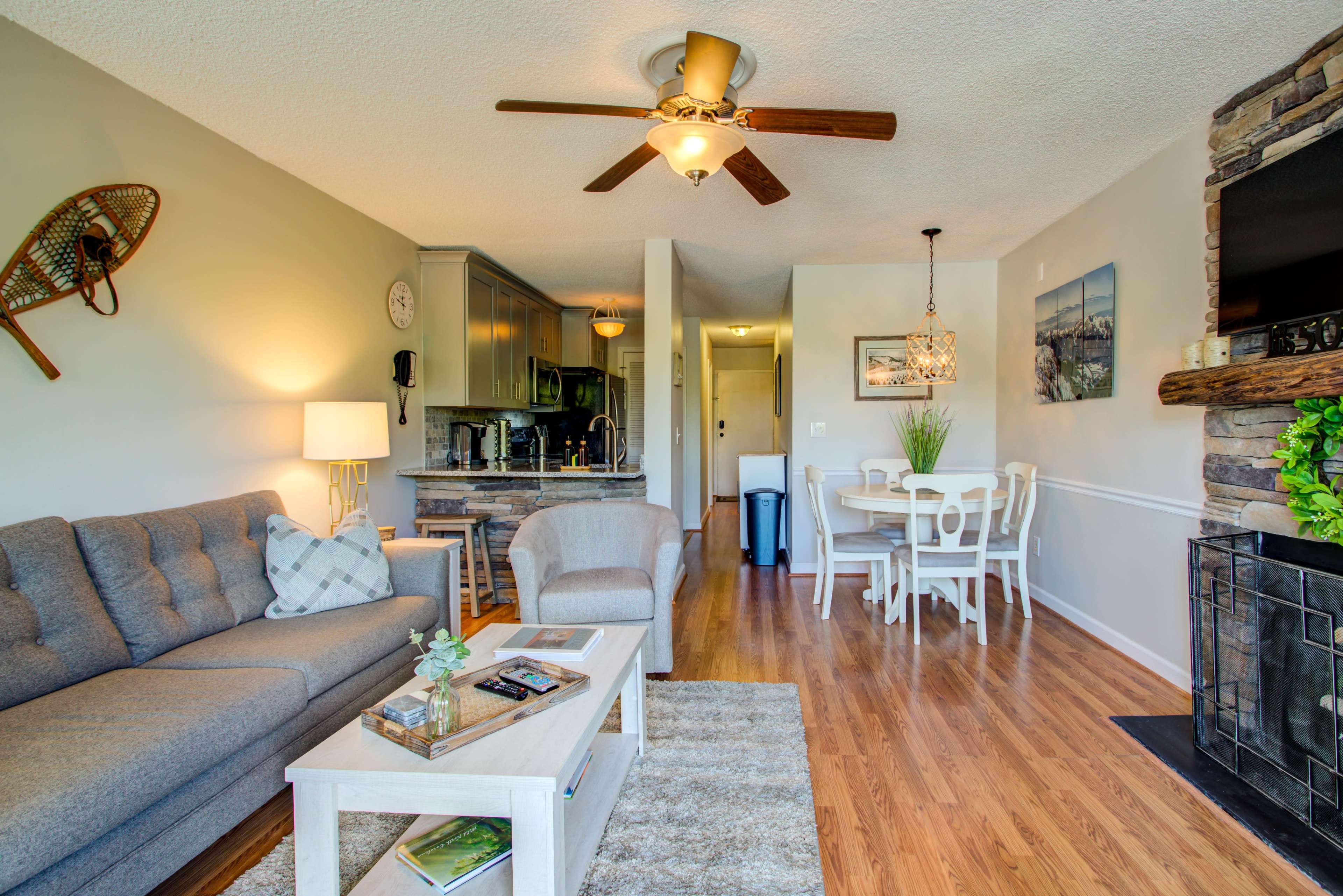 A living room features a gray sofa, a wooden coffee table, and a stone fireplace, leading into a dining area with a round table and chairs.