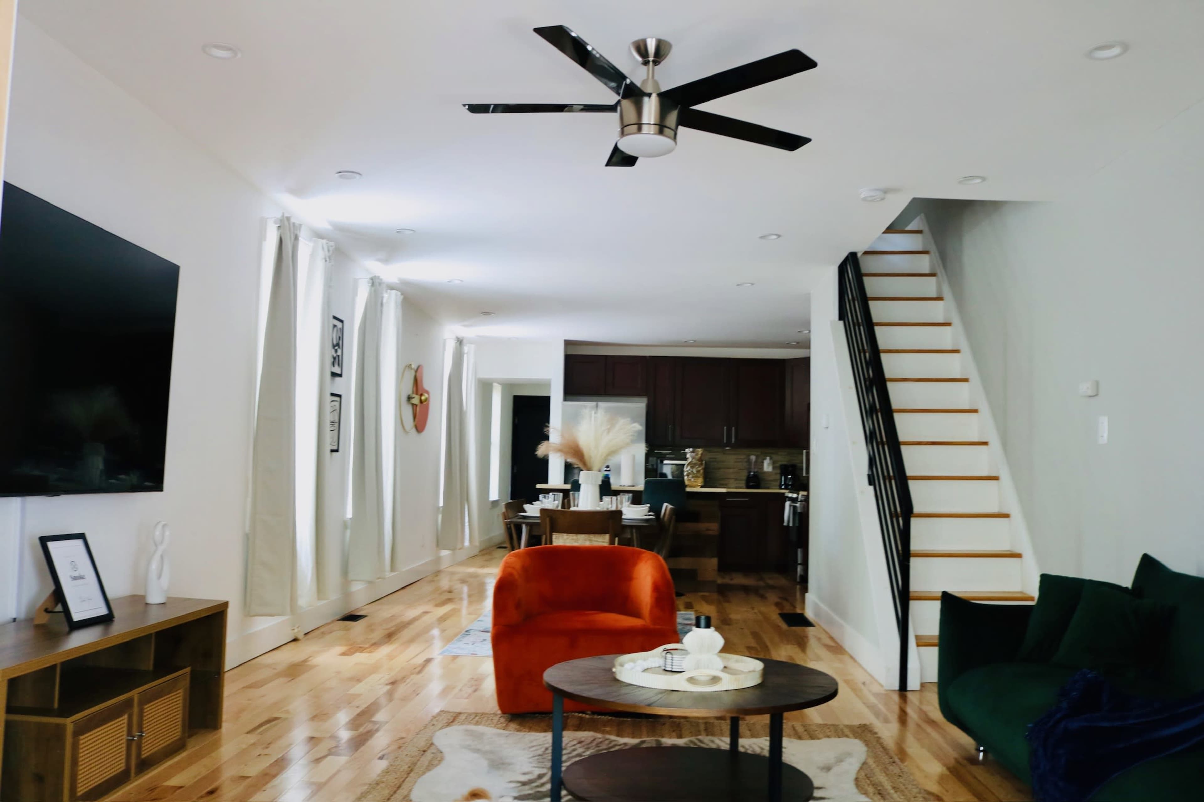 A modern living room with a staircase, featuring a kitchen in the background, a round coffee table, an orange armchair, and a green sofa.