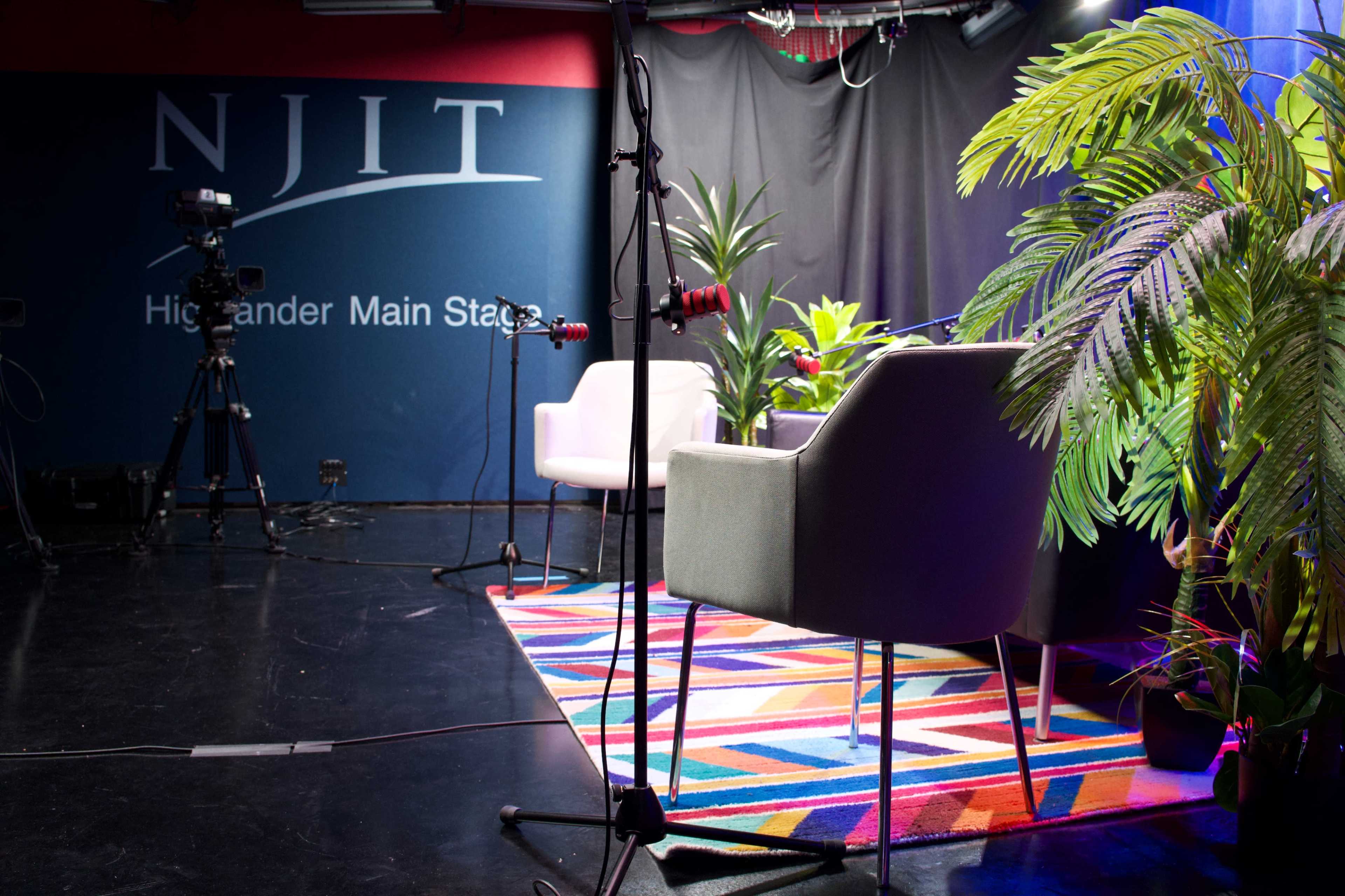 The image shows a recording studio setup featuring two chairs, a colorful rug, and a backdrop with the NJIT logo.