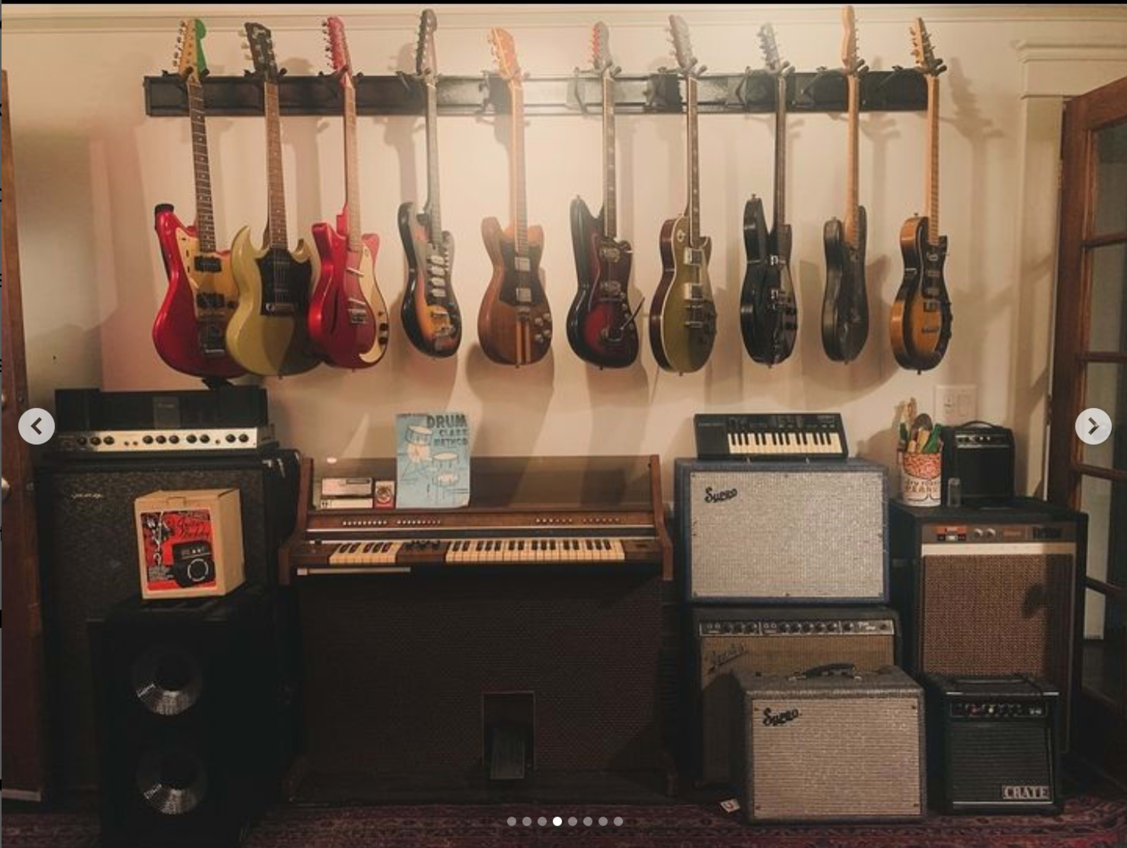 The image shows a collection of guitars hanging on a wall with various amplifiers and a vintage keyboard arranged below them.