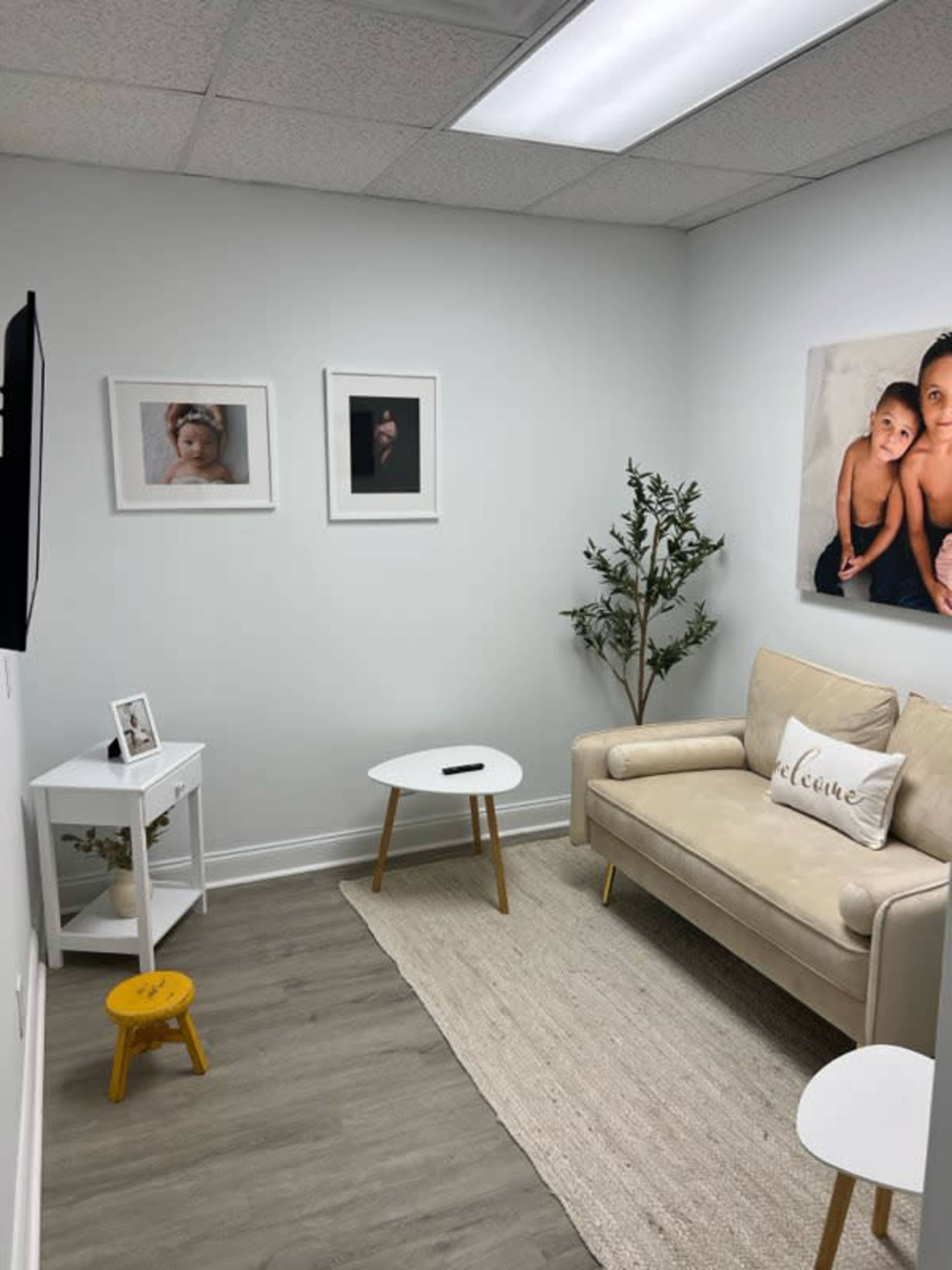 The image shows a small, minimalist living space with a beige couch, a white coffee table, and framed photographs on the wall, alongside a plant and a small yellow stool.