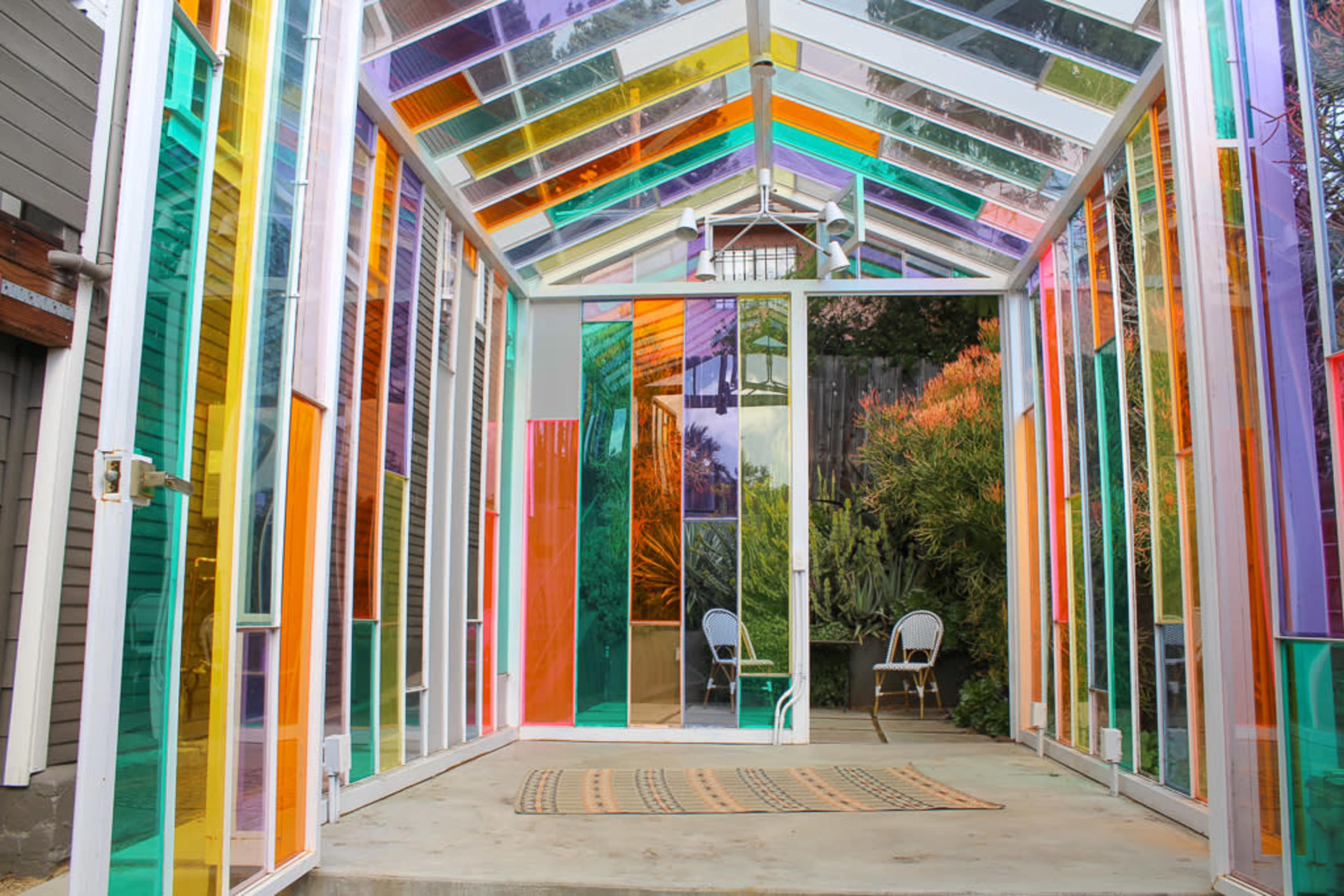 The image shows a colorful glass-walled greenhouse with varying shades of transparent panels and a small seating area at the back.