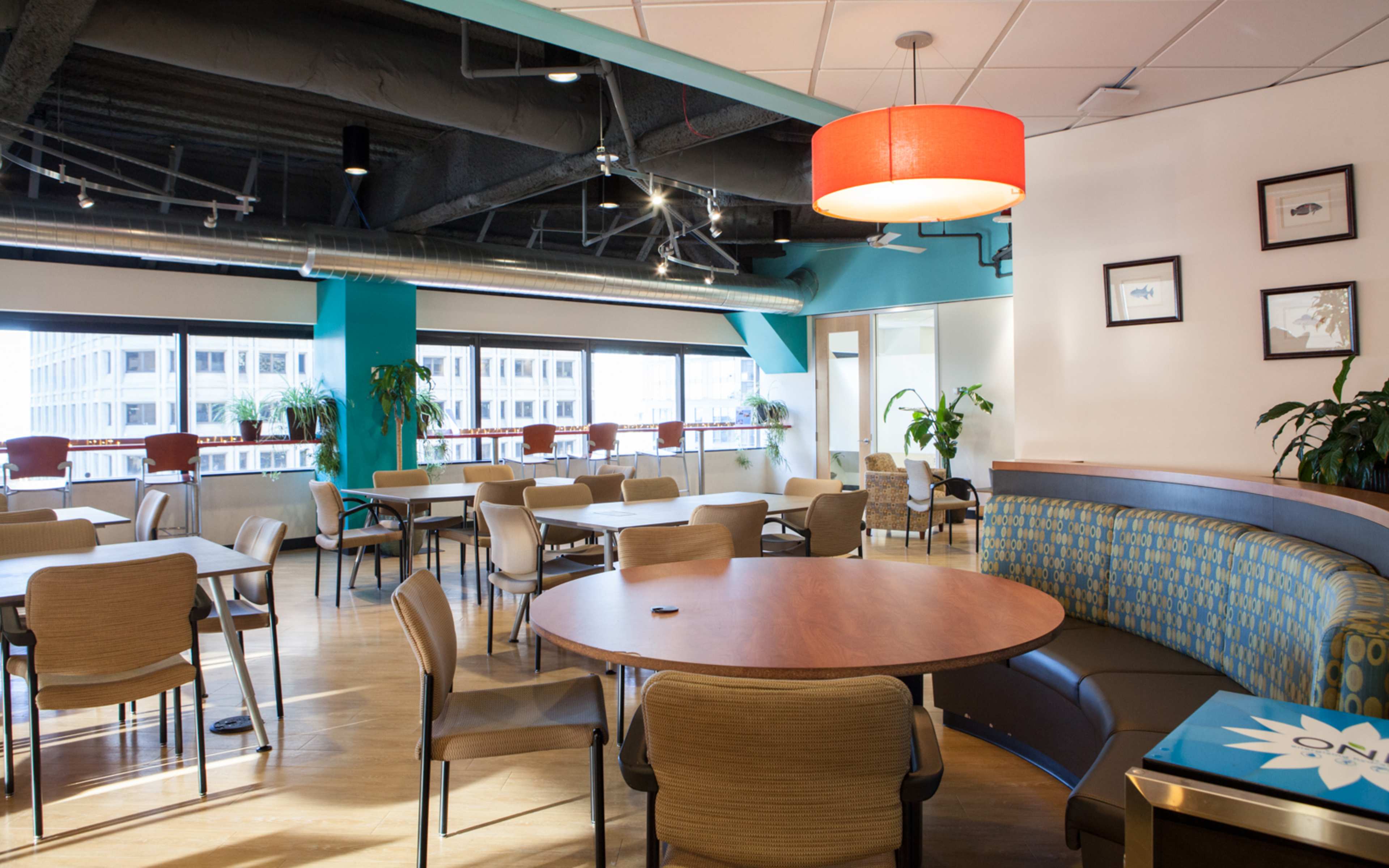 A well-lit dining area features tables and chairs arranged around circular and rectangular configurations, with large windows displaying an urban backdrop.