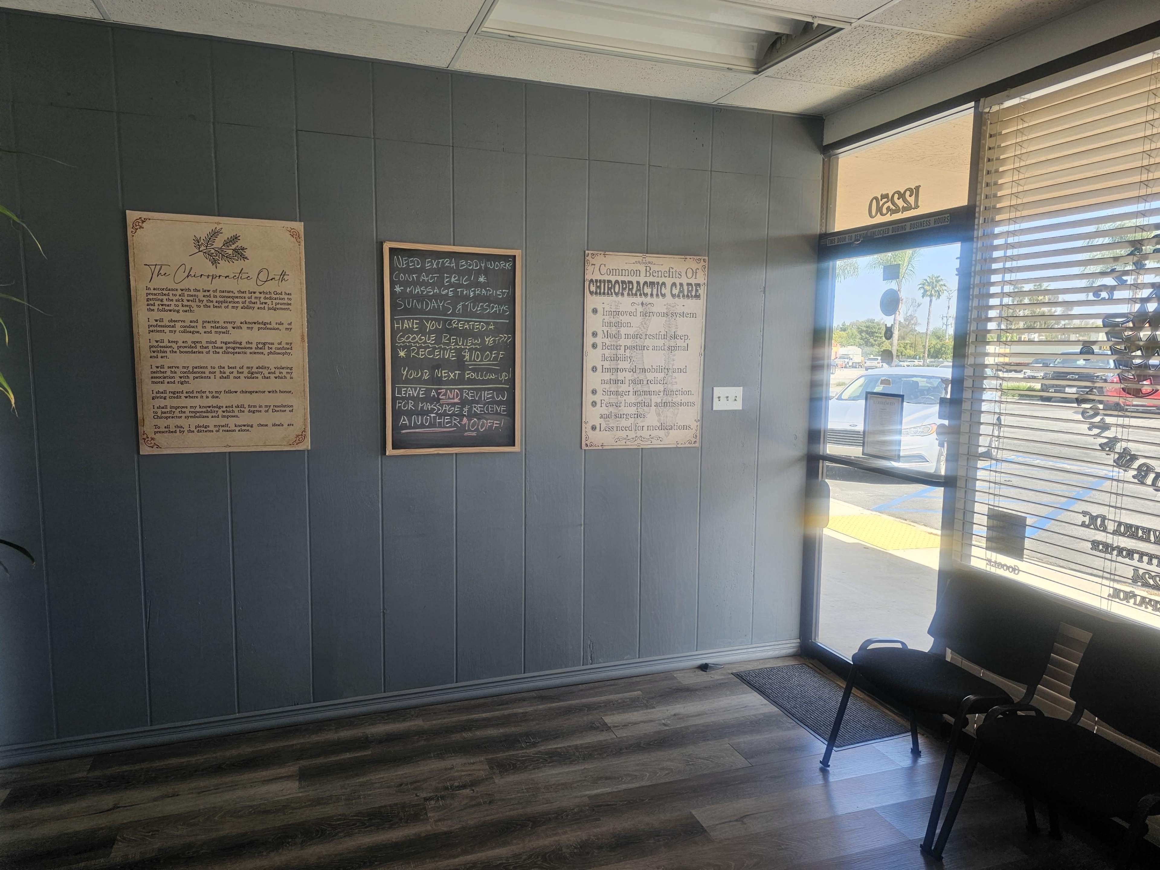 The image shows an interior waiting area with a wall featuring informational posters about chiropractic care and a large entrance door with vertical blinds.