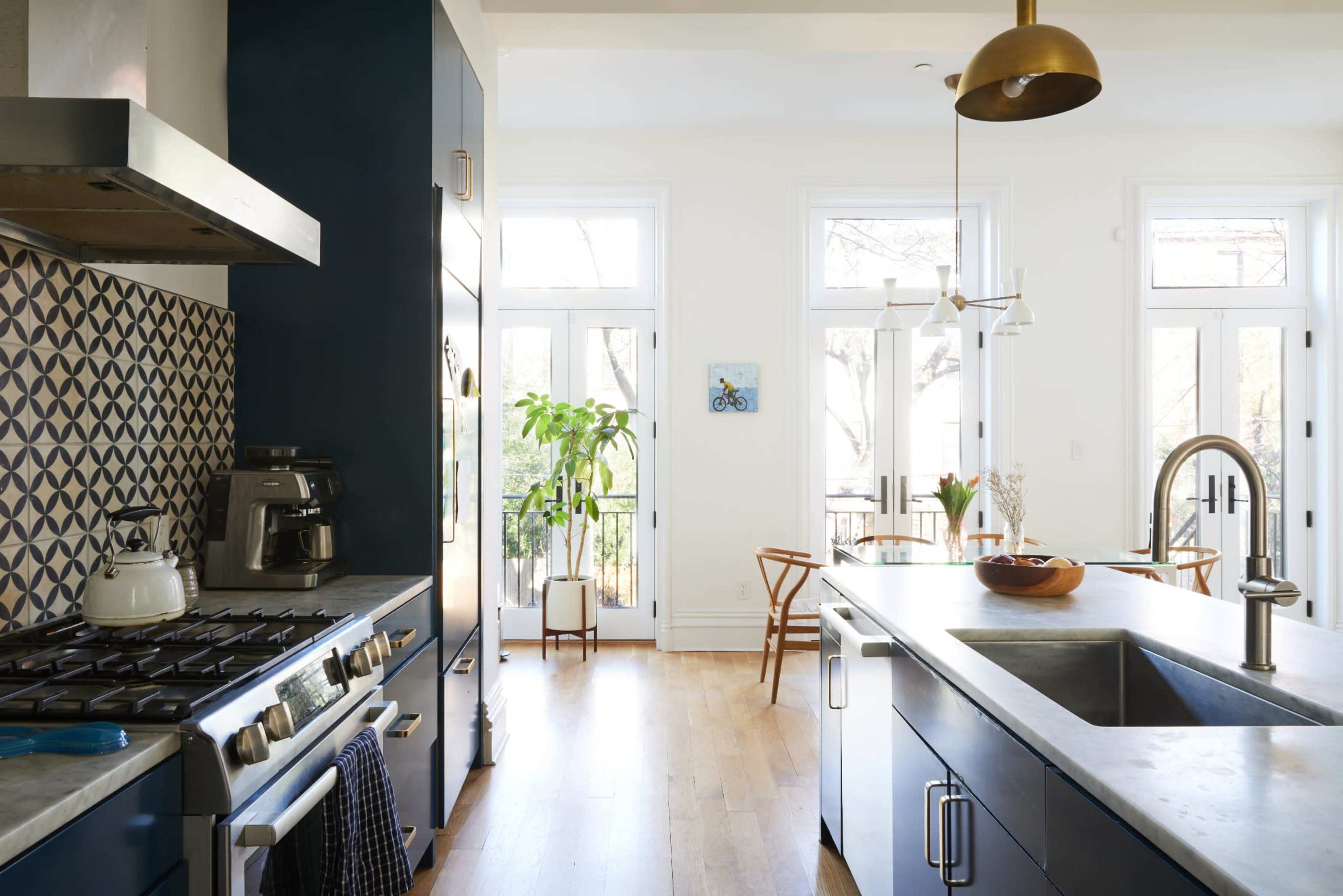 A modern kitchen features dark blue cabinetry, stainless steel appliances, and a large island with a sink, connected to a dining area with wooden chairs and plants.