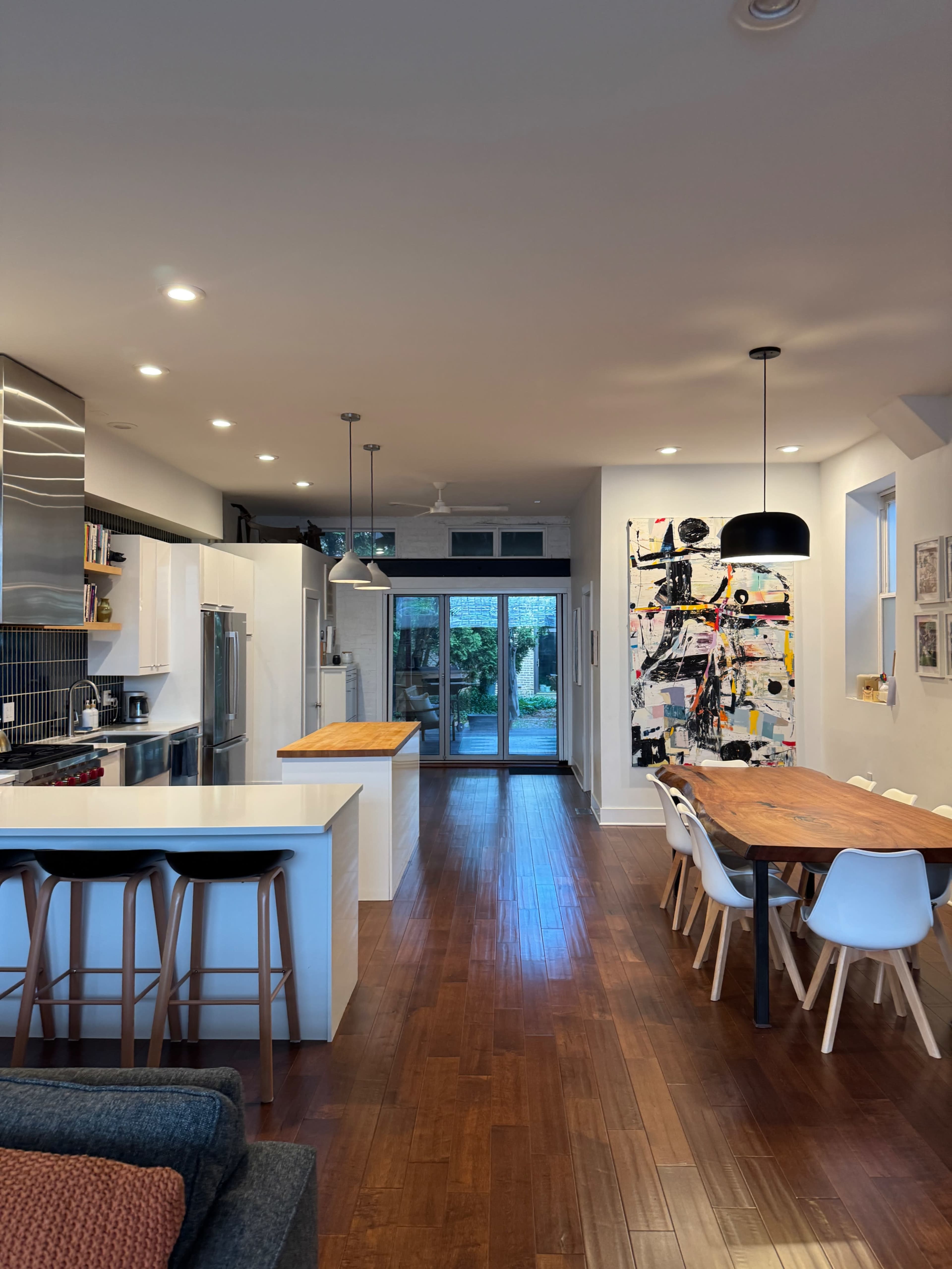 The image shows a modern kitchen and dining area with hardwood floors, featuring a large table, white cabinets, and sliding glass doors leading to a garden.