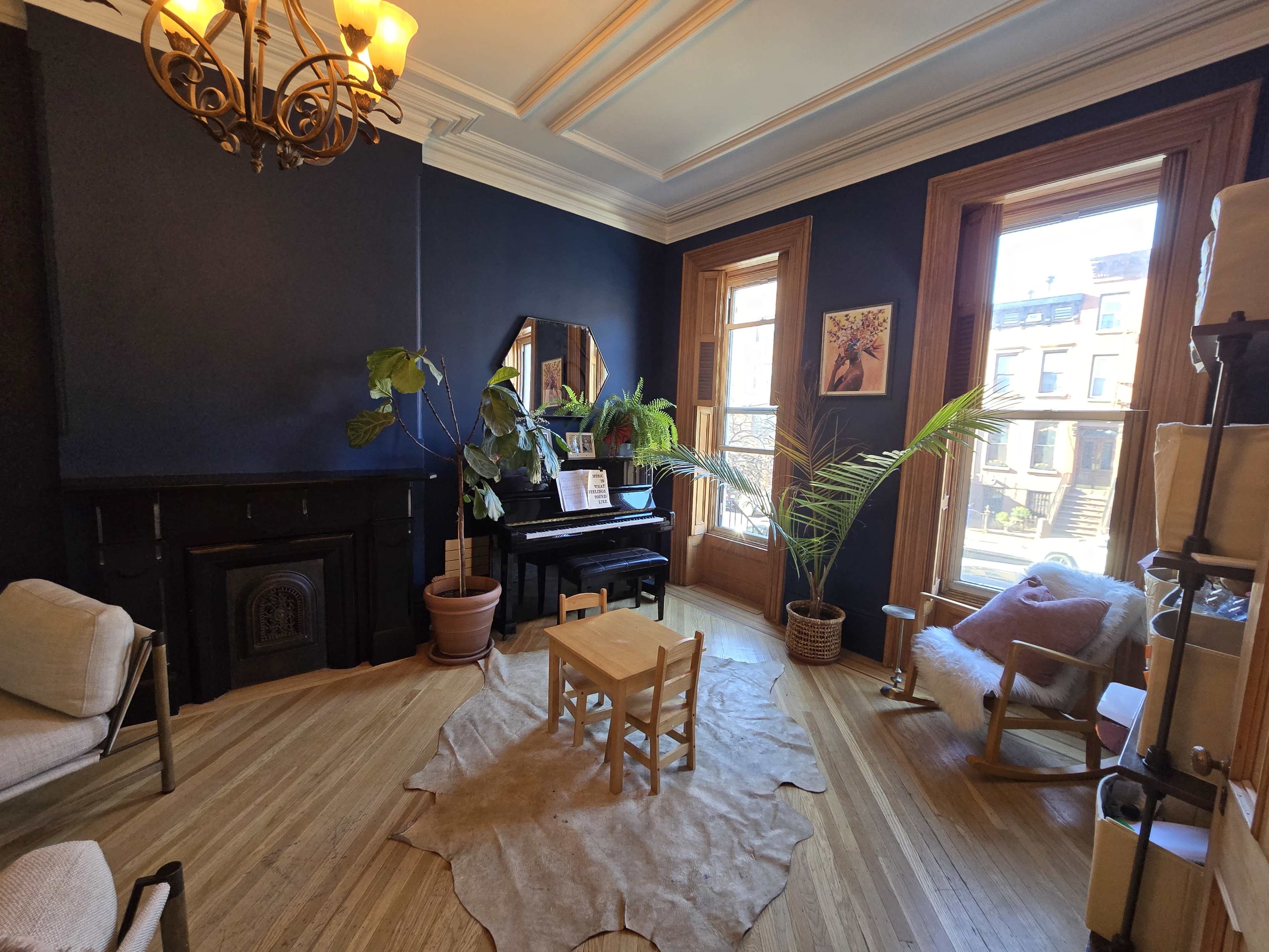 The image shows a cozy room featuring dark blue walls, a piano, wooden furniture, and natural light coming through two windows.