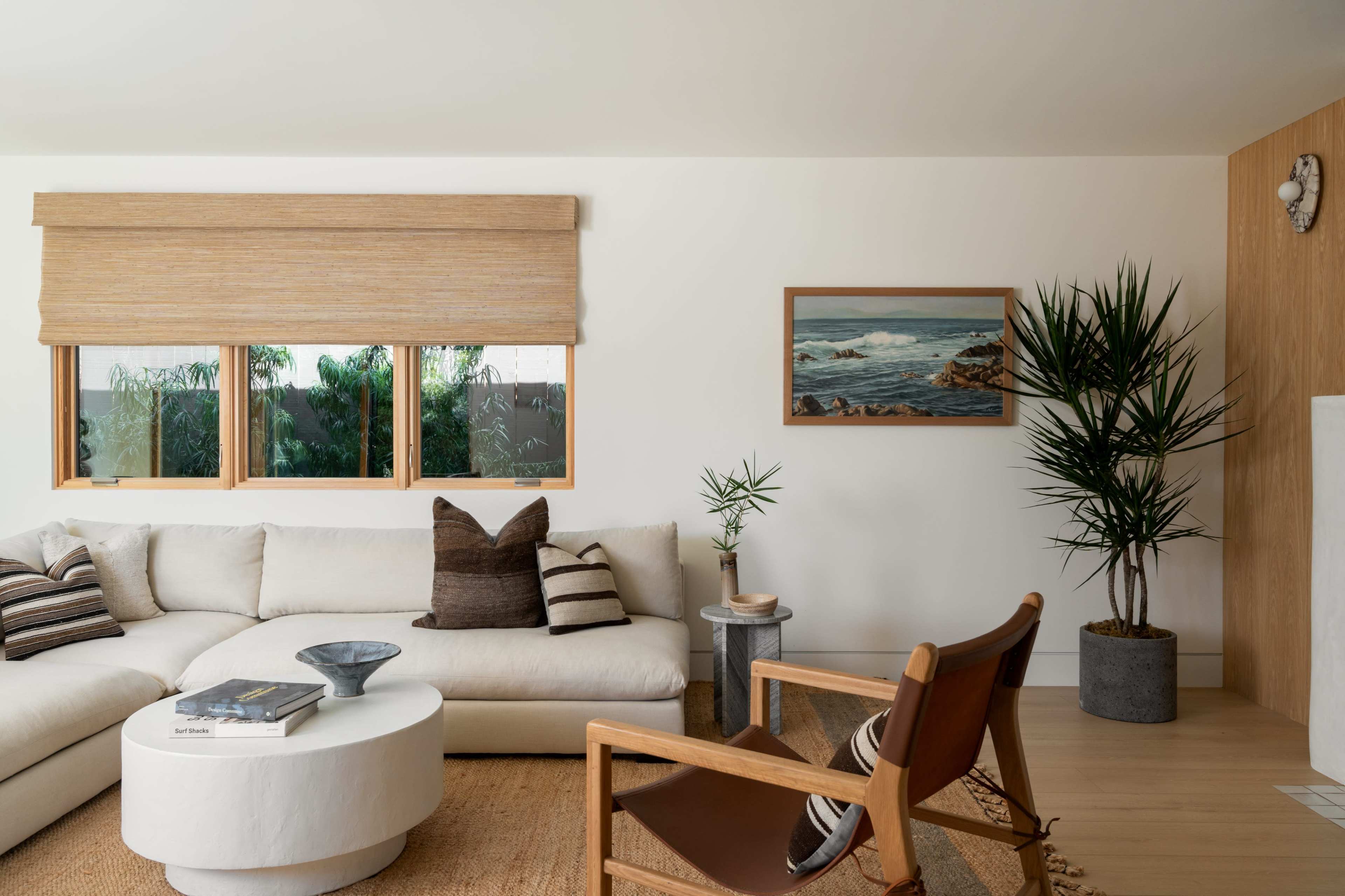 The image features a modern living room with a large white sectional sofa, a round coffee table, and a wooden chair, complemented by a tropical plant and a landscape painting on the wall.