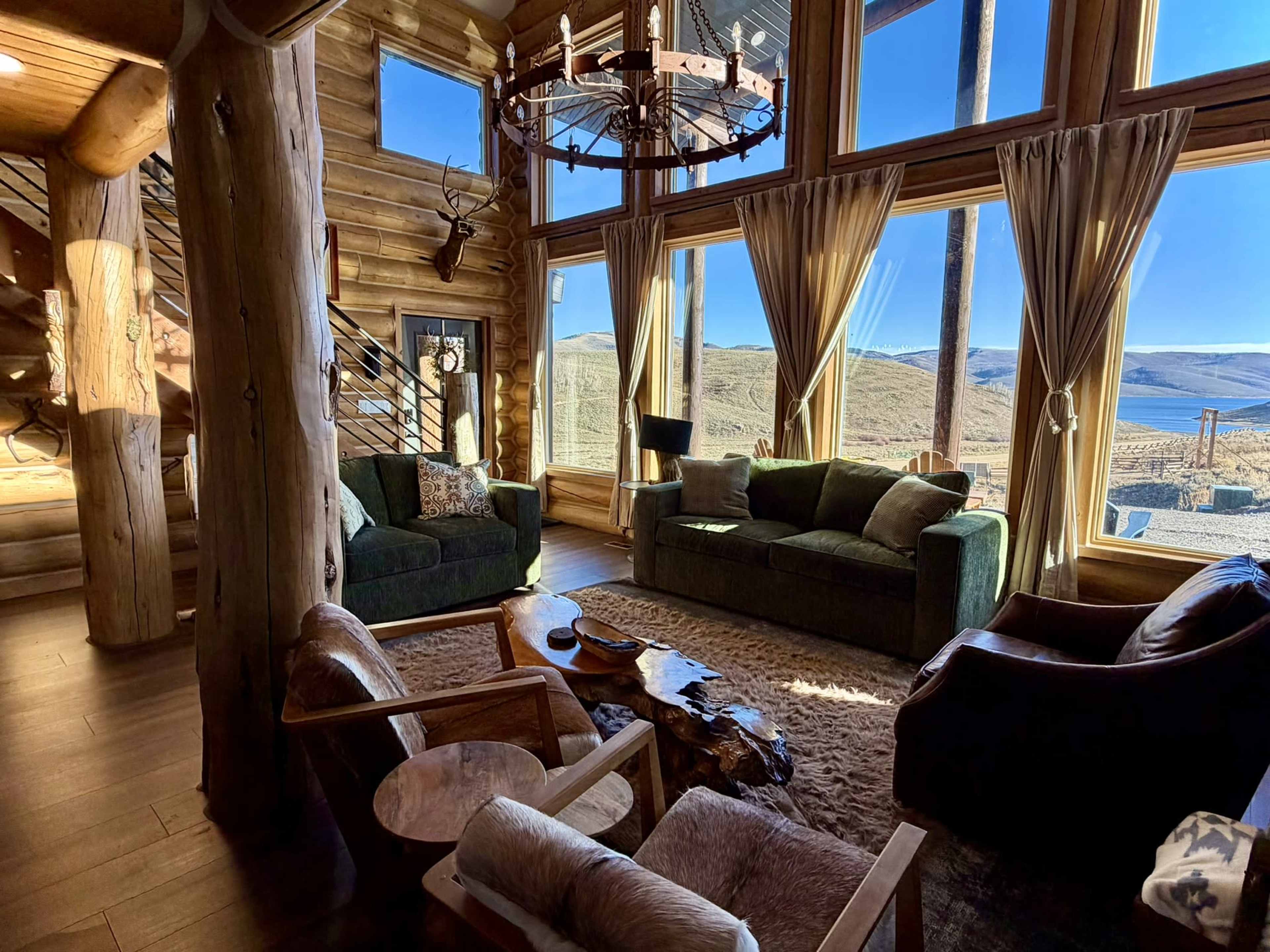 The image shows a cozy log cabin interior with large windows, offering a view of a mountainous landscape, and features a seating area with various couches and a wooden coffee table.