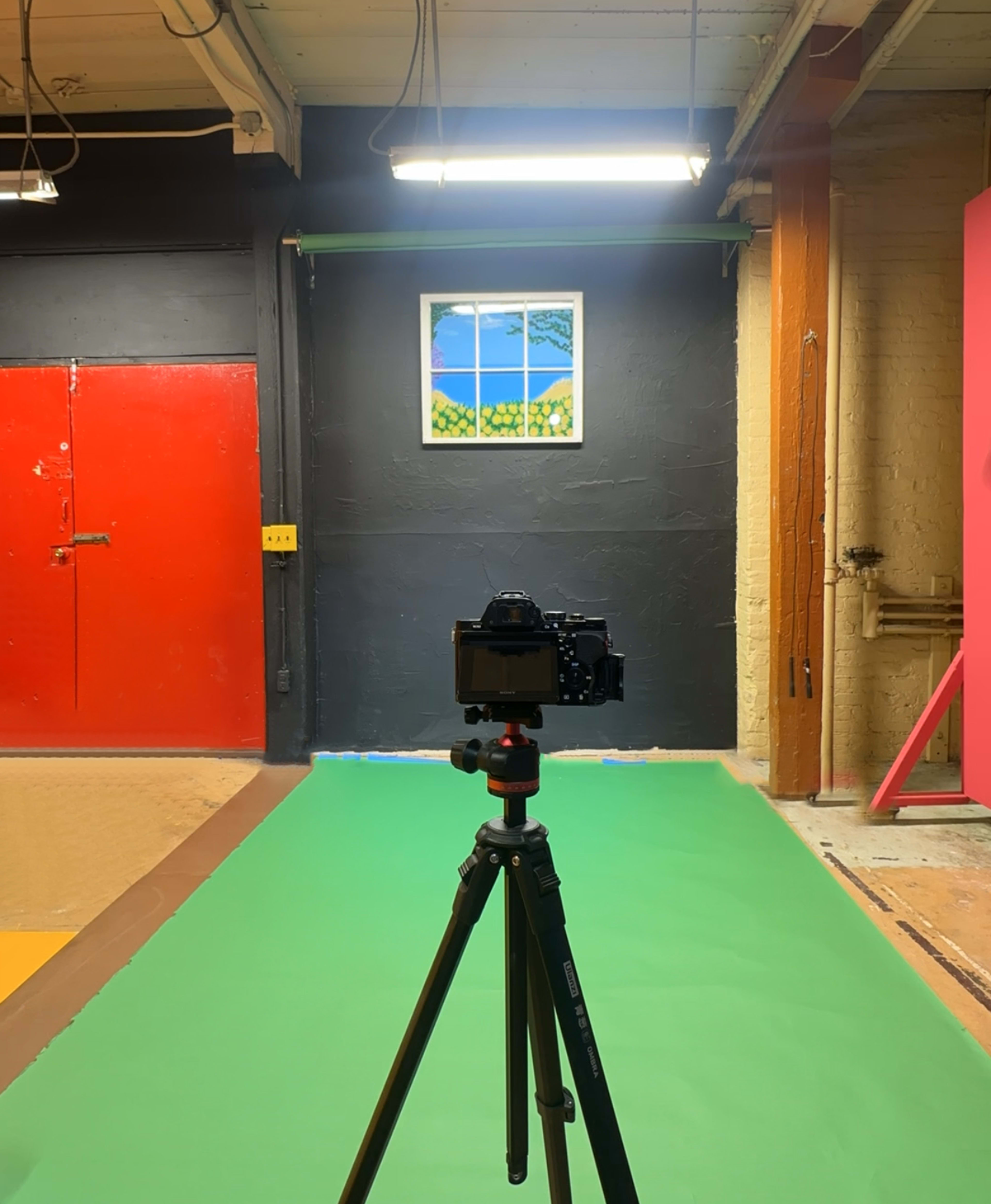 A camera mounted on a tripod is positioned in front of a mural depicting a window with a landscape, against a backdrop of colorful walls in a studio.