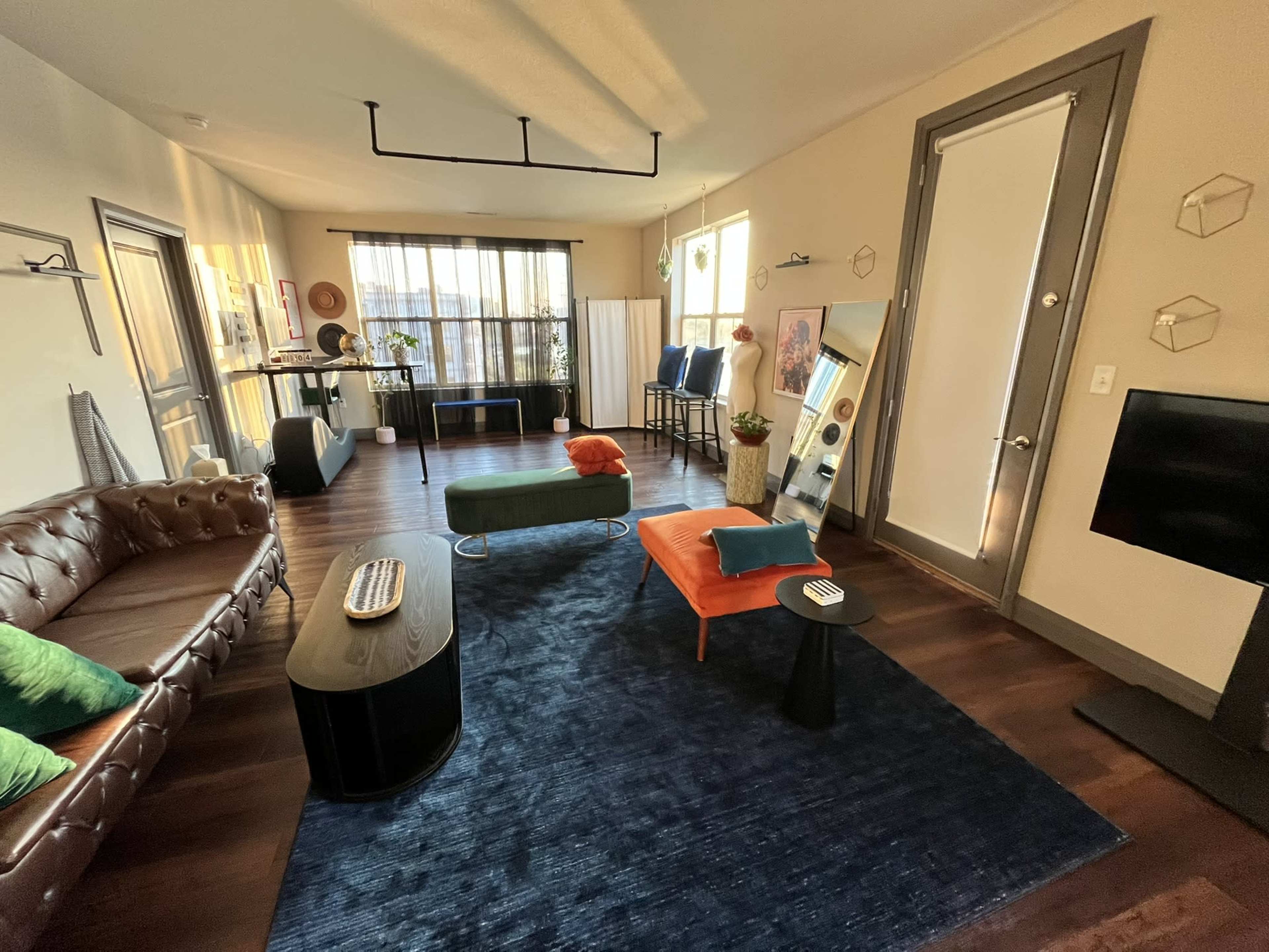 The image shows a modern living room featuring a brown leather sofa, a green and orange upholstered chair, a dark coffee table, and a large window letting in natural light.